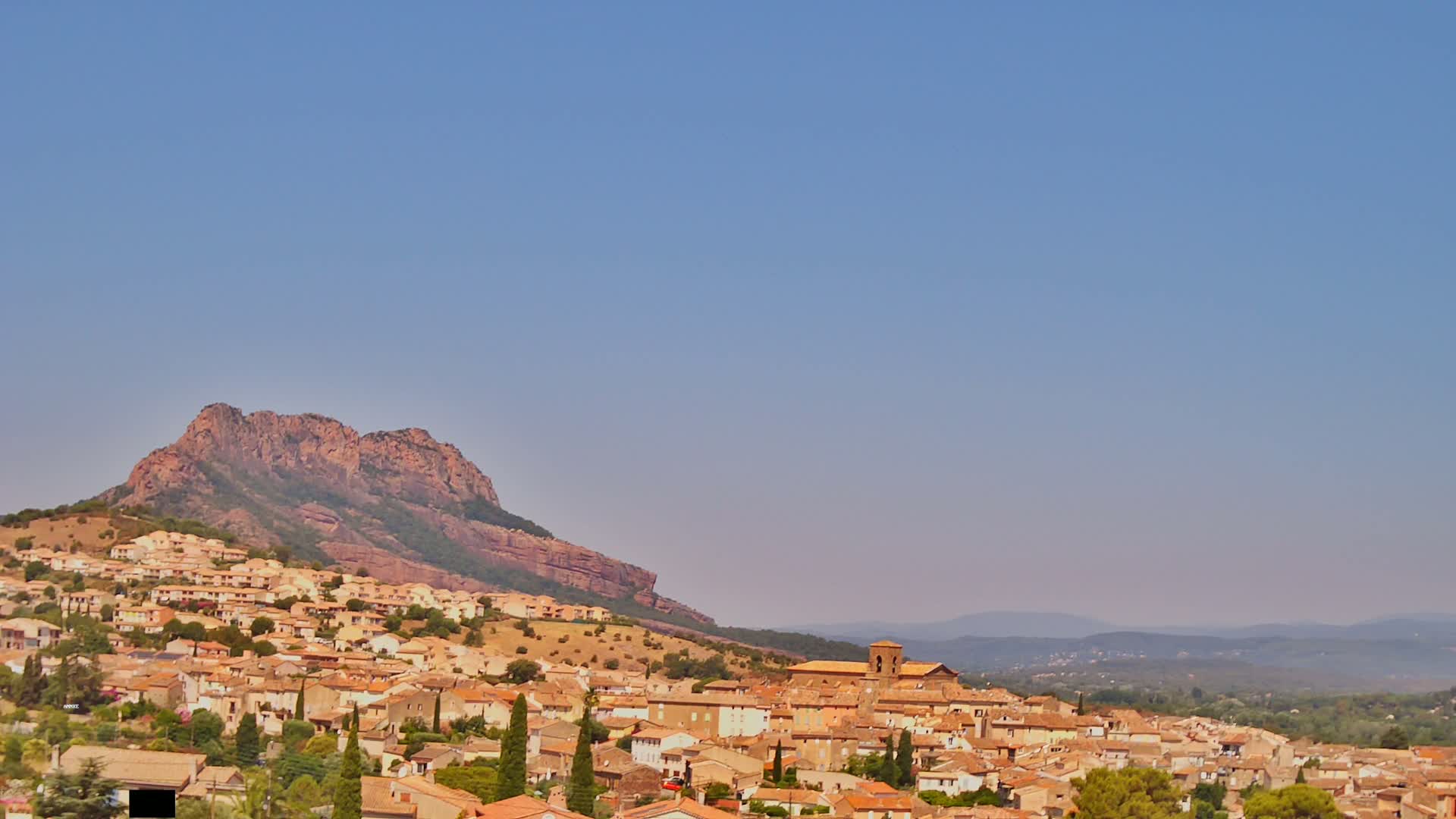 A small town of tan and brown buildings nestled against a large, reddish-brown mountain under a clear, pale blue sky.