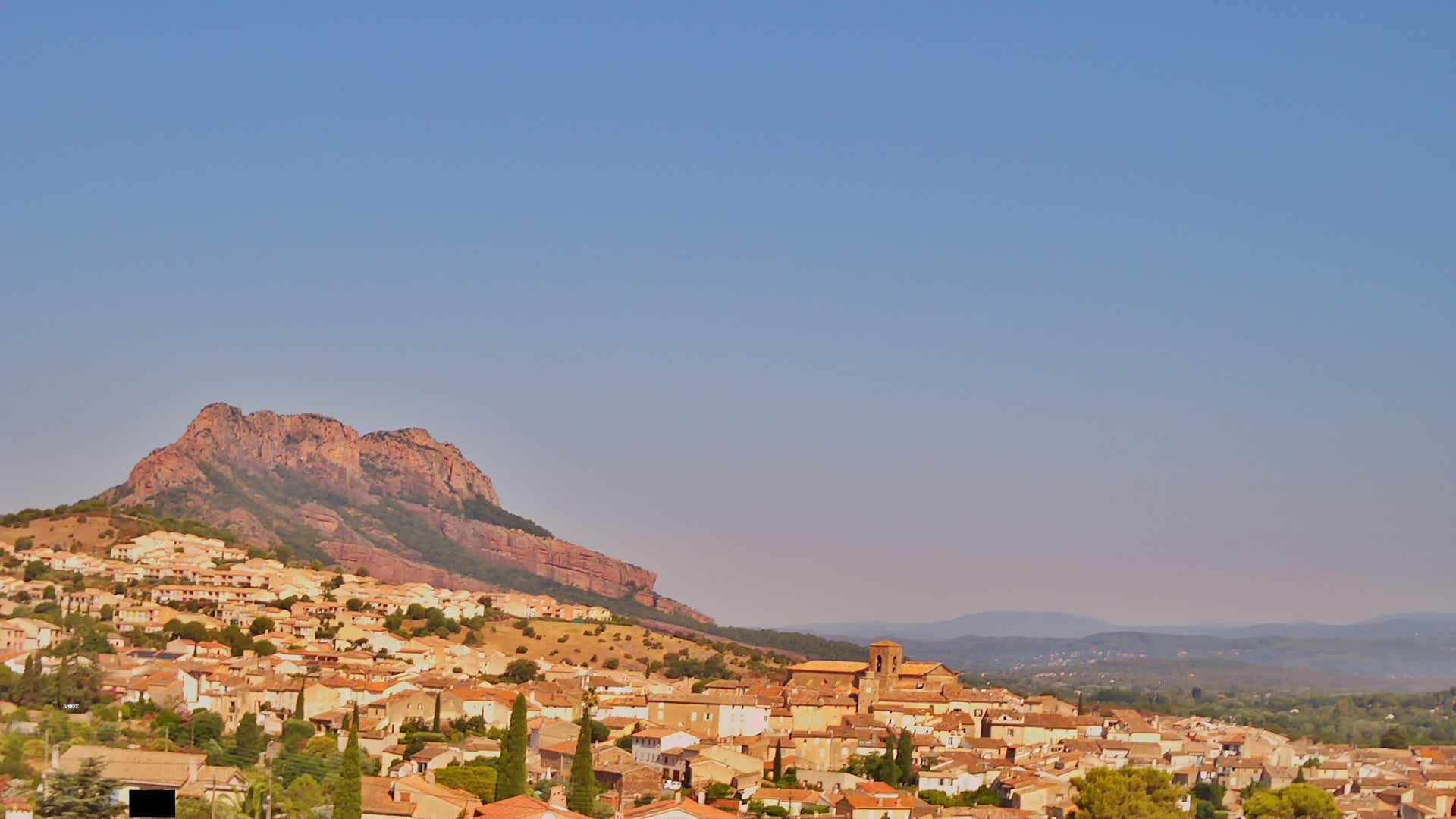 A small town with tan-colored buildings nestled against a large reddish-brown hill under a clear, light-blue sky.