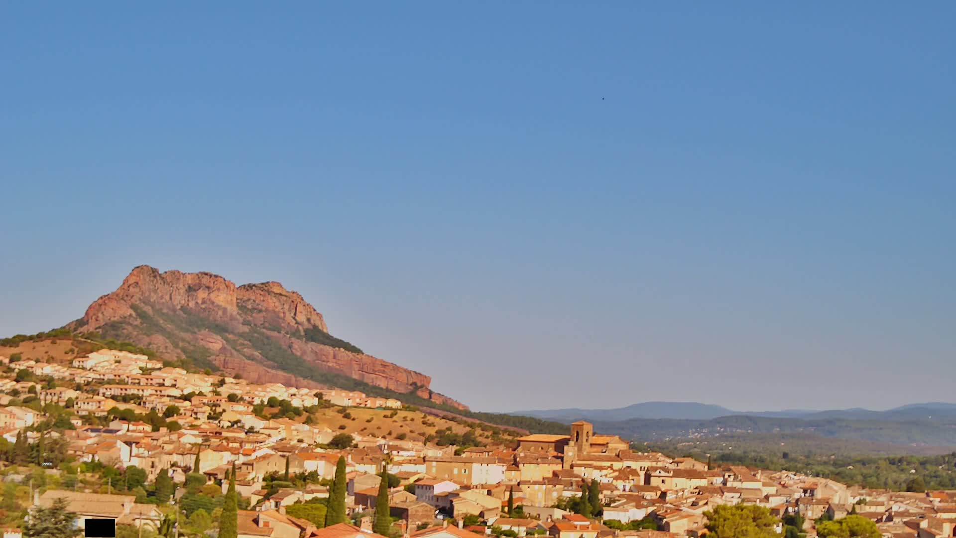 A tan-colored village sits nestled at the base of a large, reddish-brown mountain under a clear, blue sky.