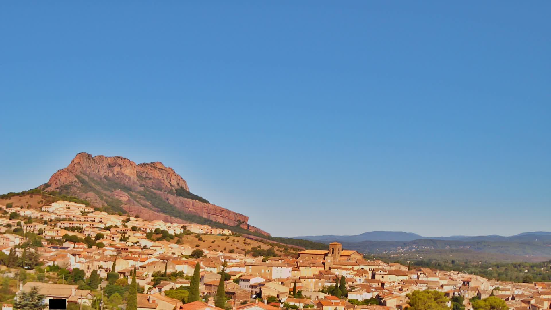 A village of tan and brown buildings sprawls across a hillside beneath a large, reddish-brown rock formation, all under a clear blue sky.