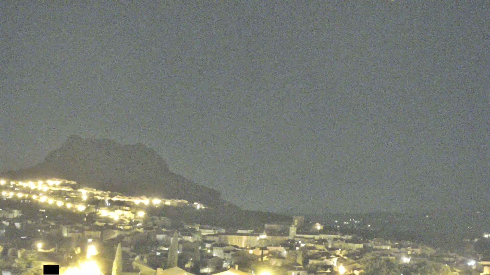 A nighttime view shows a hilltop town illuminated by lights, with a dark mountain rising behind it under a cloudy sky.