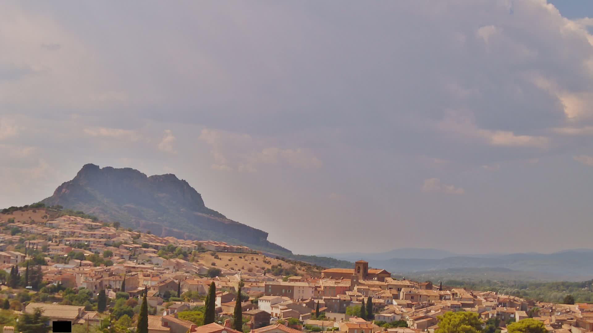 A nighttime view shows a hilltop town illuminated by lights, with a dark mountain rising behind it under a cloudy sky.