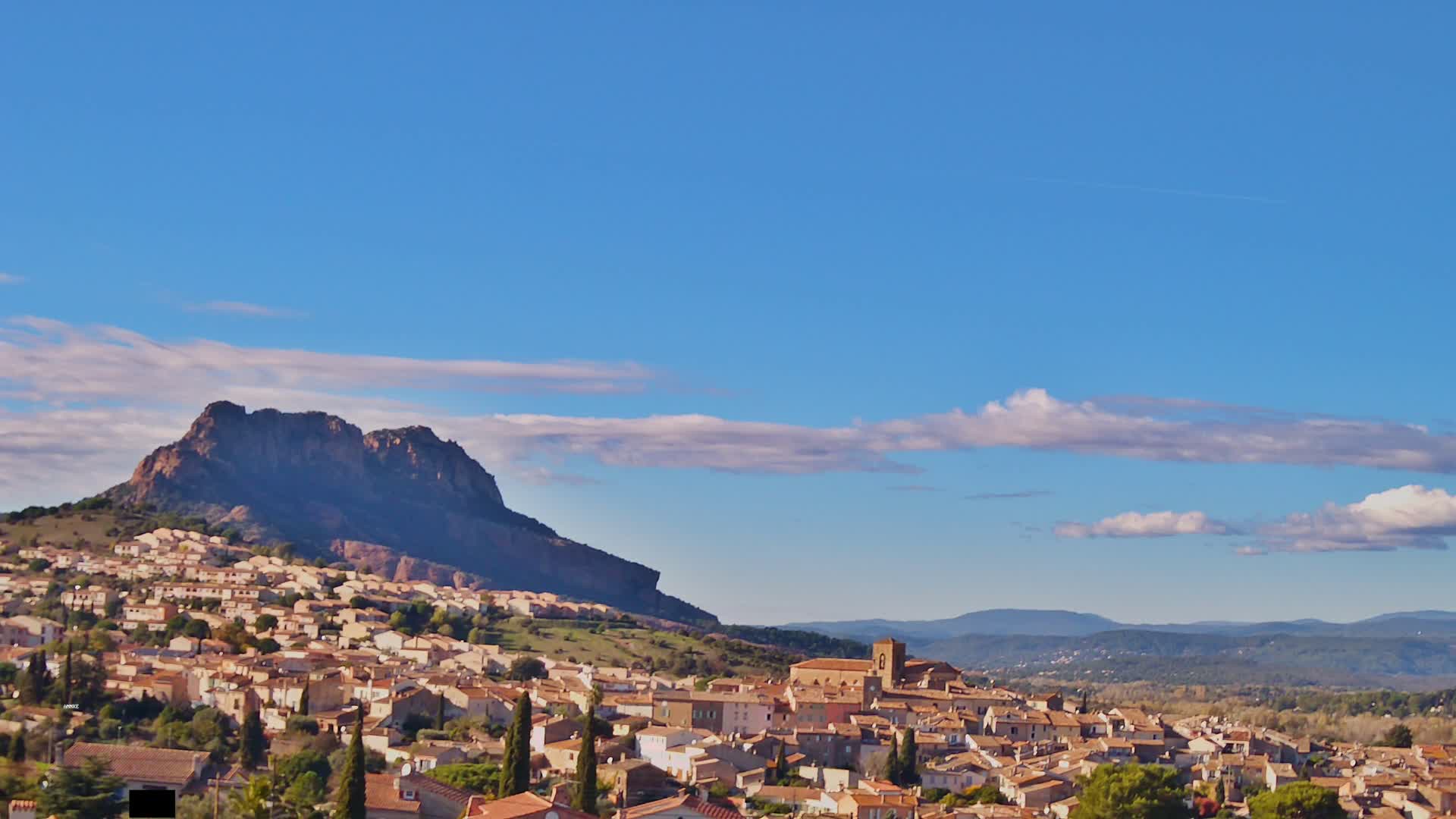 A nighttime view shows a hilltop town illuminated by lights, with a dark mountain rising behind it under a cloudy sky.