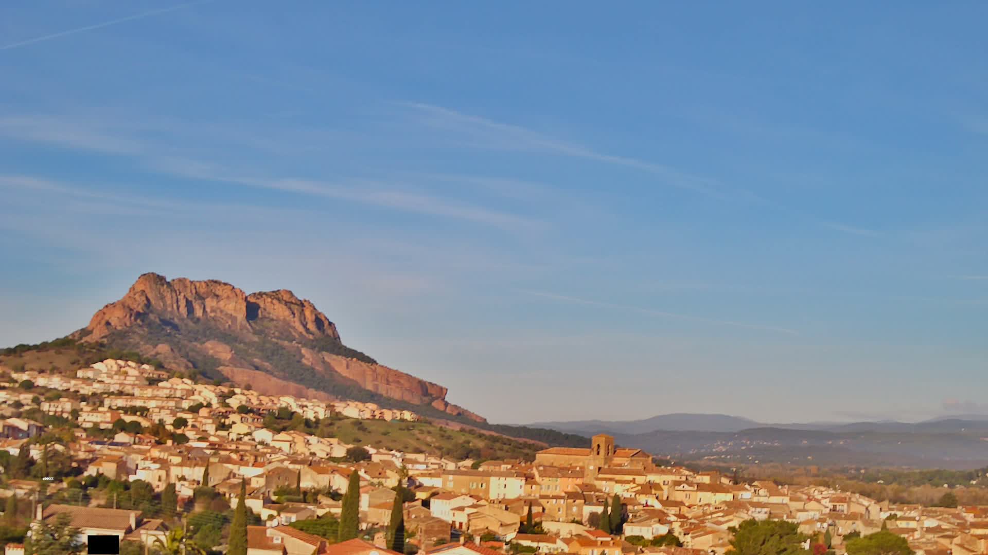 A picturesque village with numerous terracotta-roofed buildings is nestled against the base of a large, rugged reddish-brown mountain, all bathed in sunlight under a clear blue sky with faint contrails.
