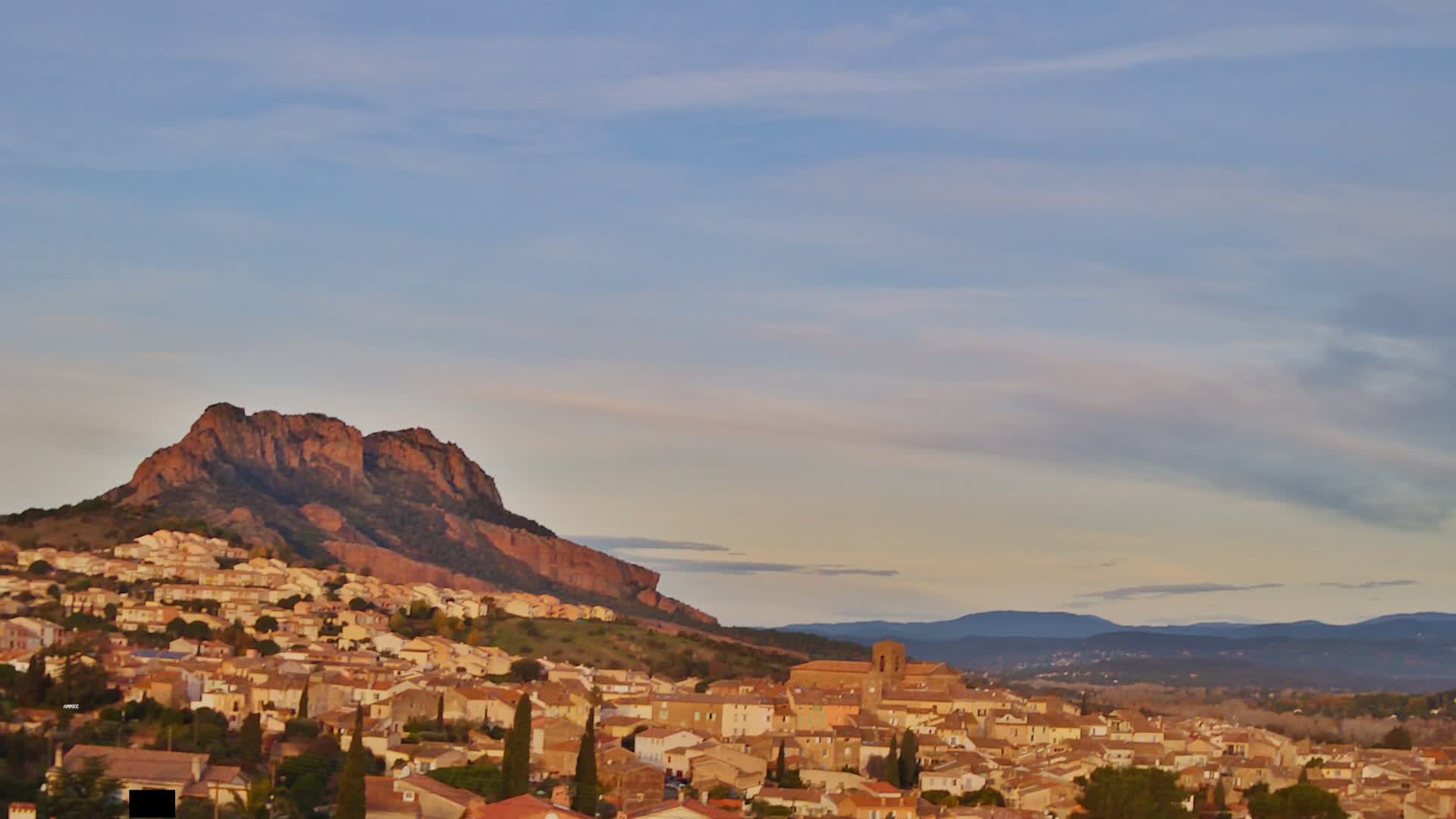 A picturesque town with light-colored buildings and a prominent church is nestled at the base of a large, reddish rocky mountain, all under a sky with scattered clouds bathed in warm golden hour light.