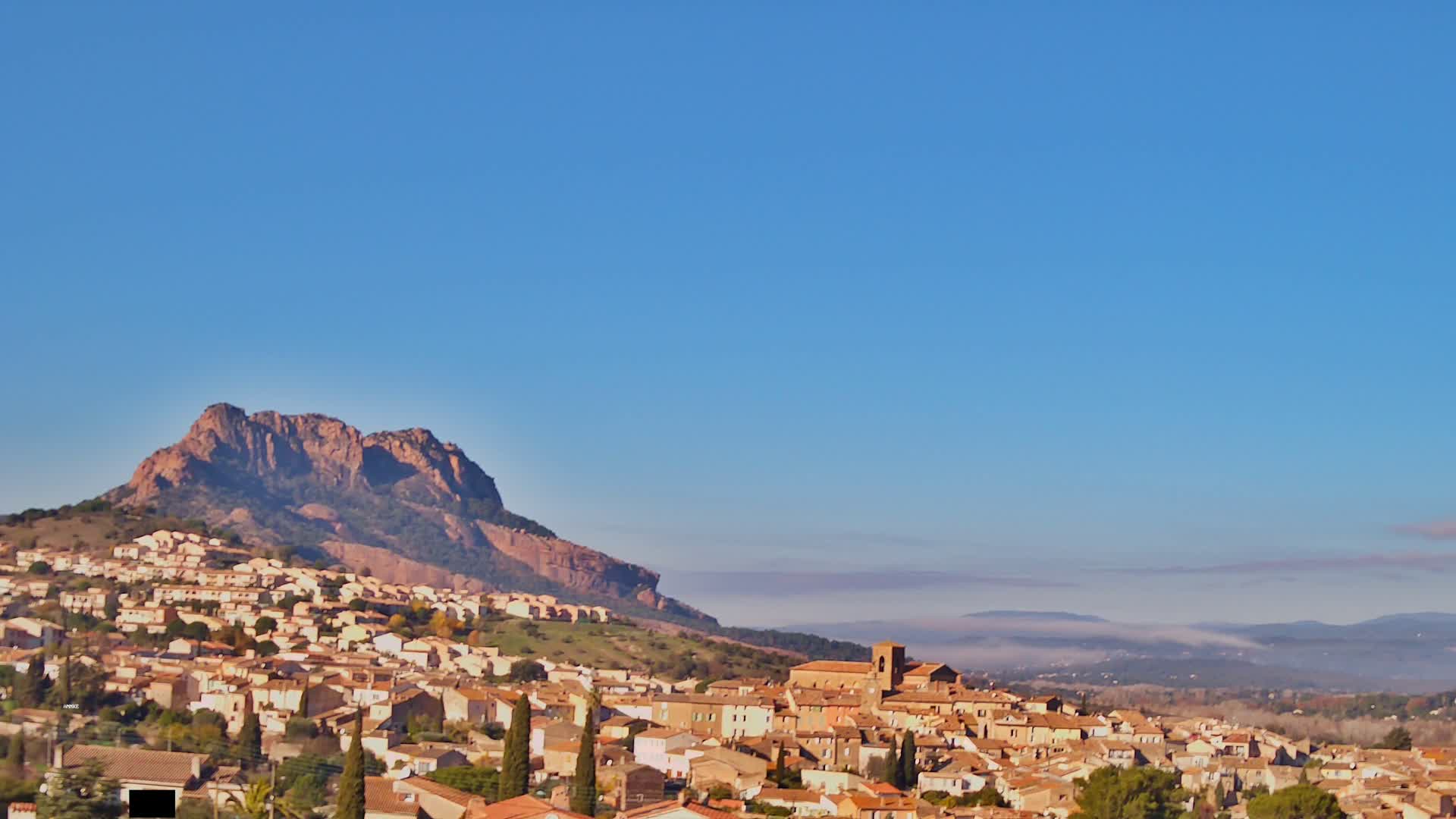 A picturesque town with light-colored buildings and a prominent church is nestled at the base of a large, reddish rocky mountain, all under a sky with scattered clouds bathed in warm golden hour light.