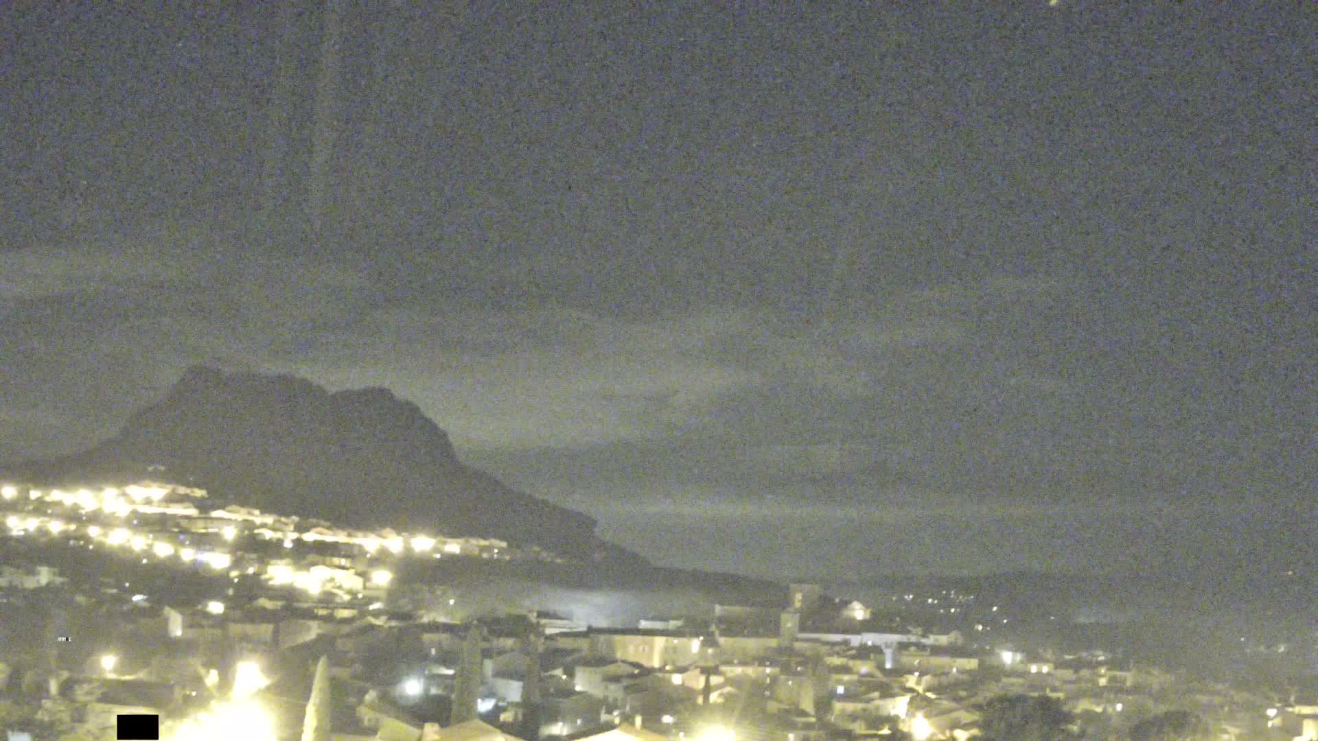 A picturesque town with light-colored buildings and a prominent church is nestled at the base of a large, reddish rocky mountain, all under a sky with scattered clouds bathed in warm golden hour light.