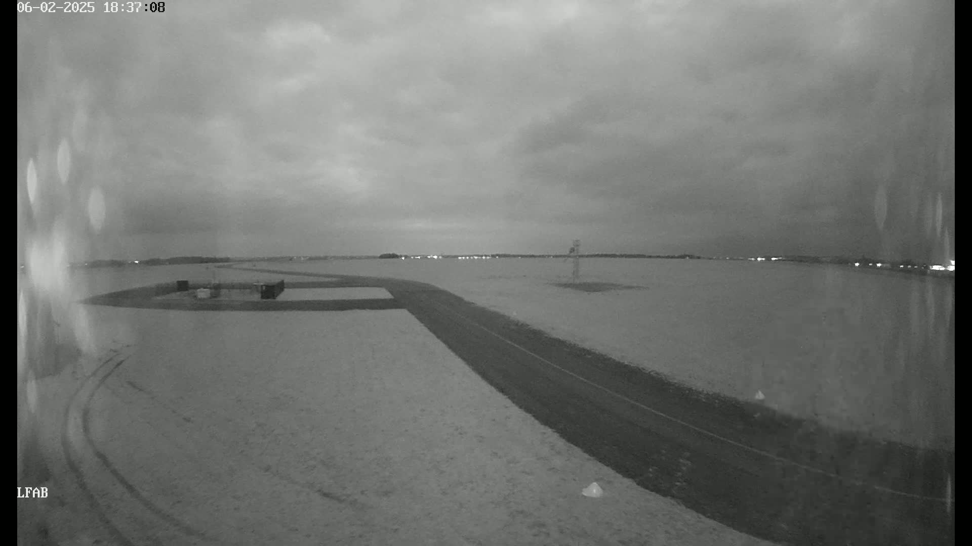 A snow-covered airfield is seen at dusk under a cloudy sky, with a paved road leading towards a distant line of lights.