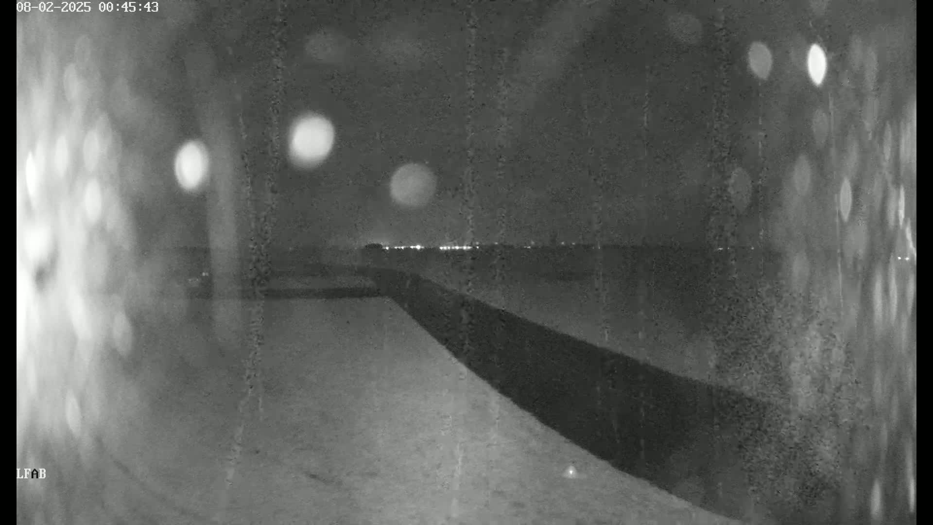 A nighttime, snowy scene shows a dark wall or embankment sloping down to a flat, snow-covered area, with distant city lights visible on the horizon.