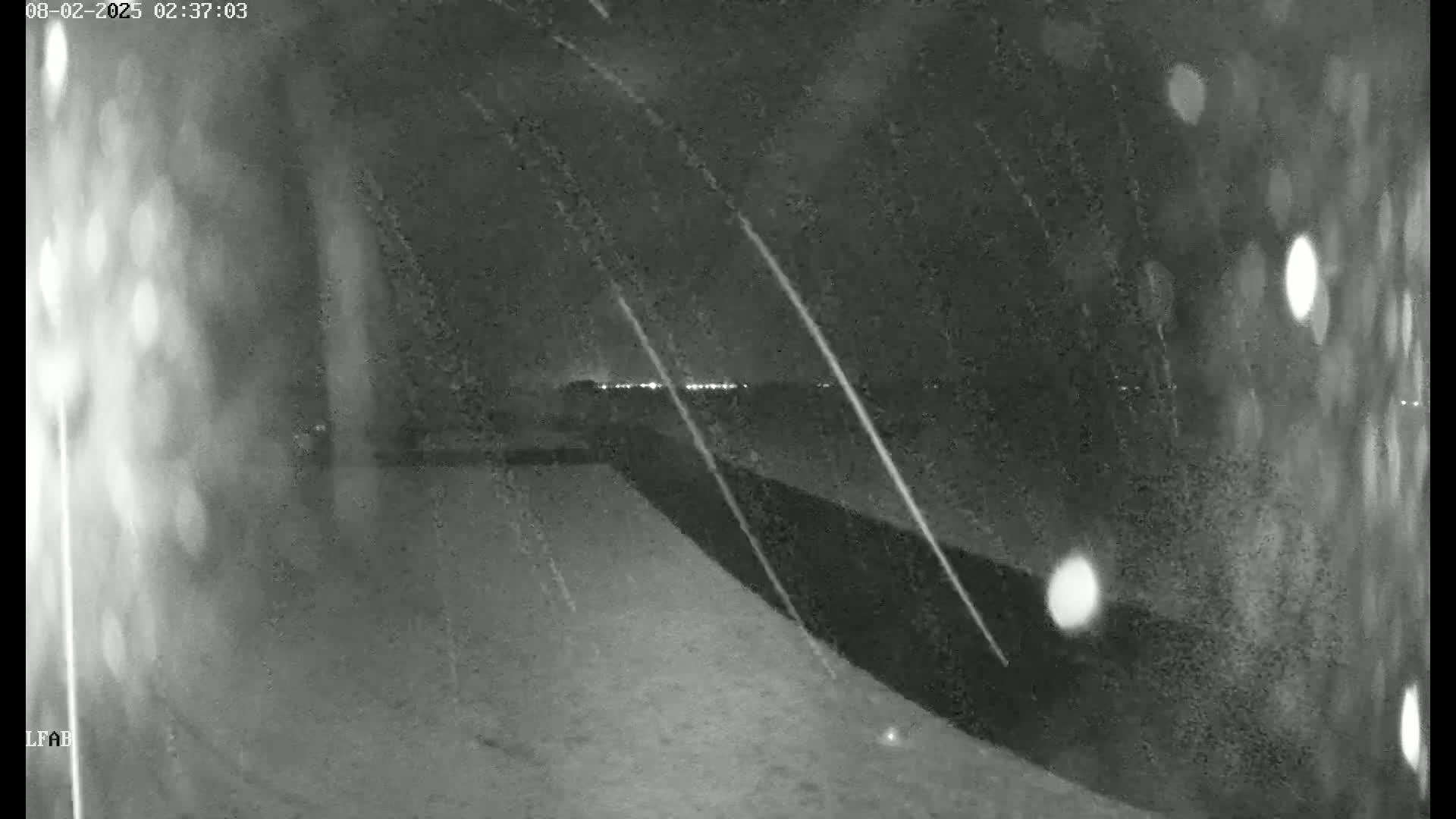 A nighttime, snowy scene shows a dark wall or fence, with faint lights in the distance.
