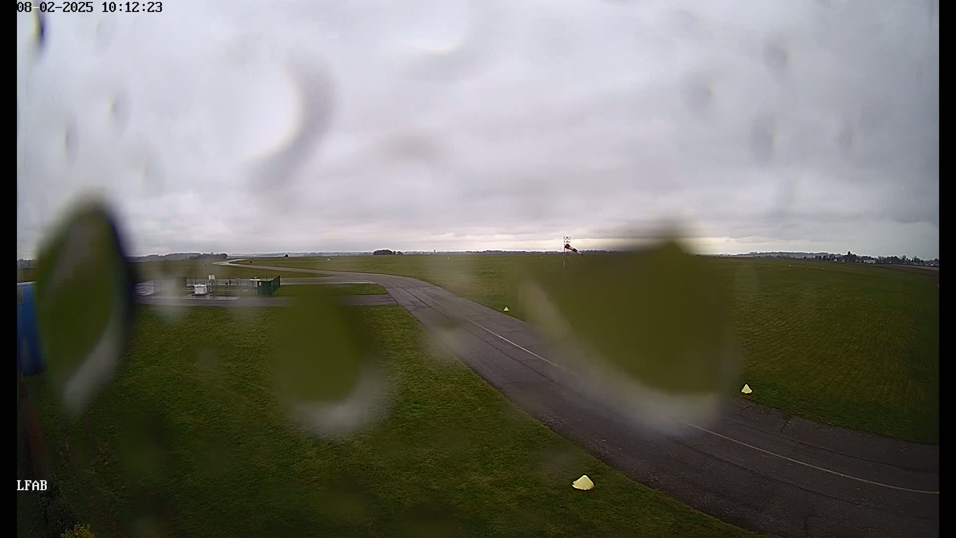 A rain-covered view of a grassy airfield with a taxiway and windsock under an overcast sky.