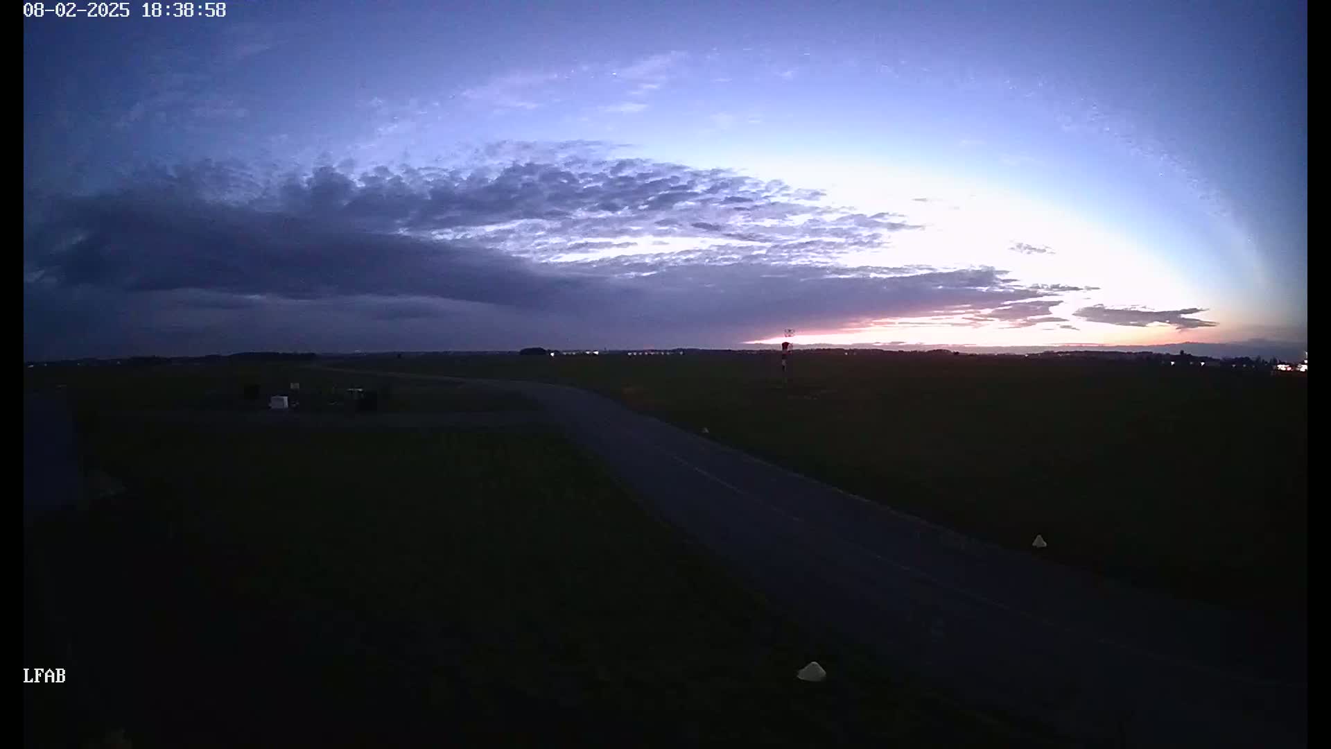 A dark road curves through a flat, grassy landscape under a twilight sky with a layer of clouds.