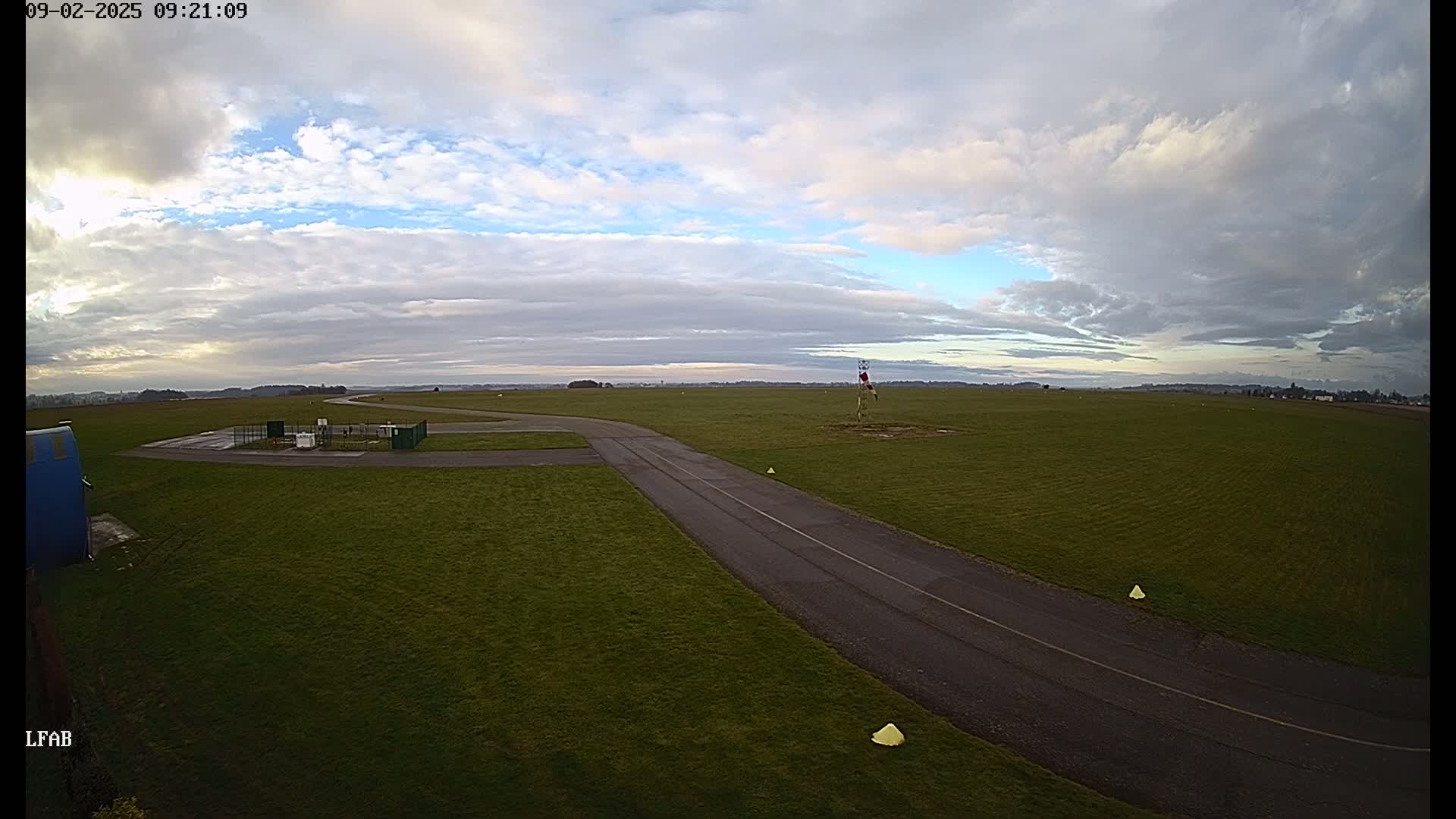 A paved taxiway curves through a grassy airfield under a partly cloudy sky.