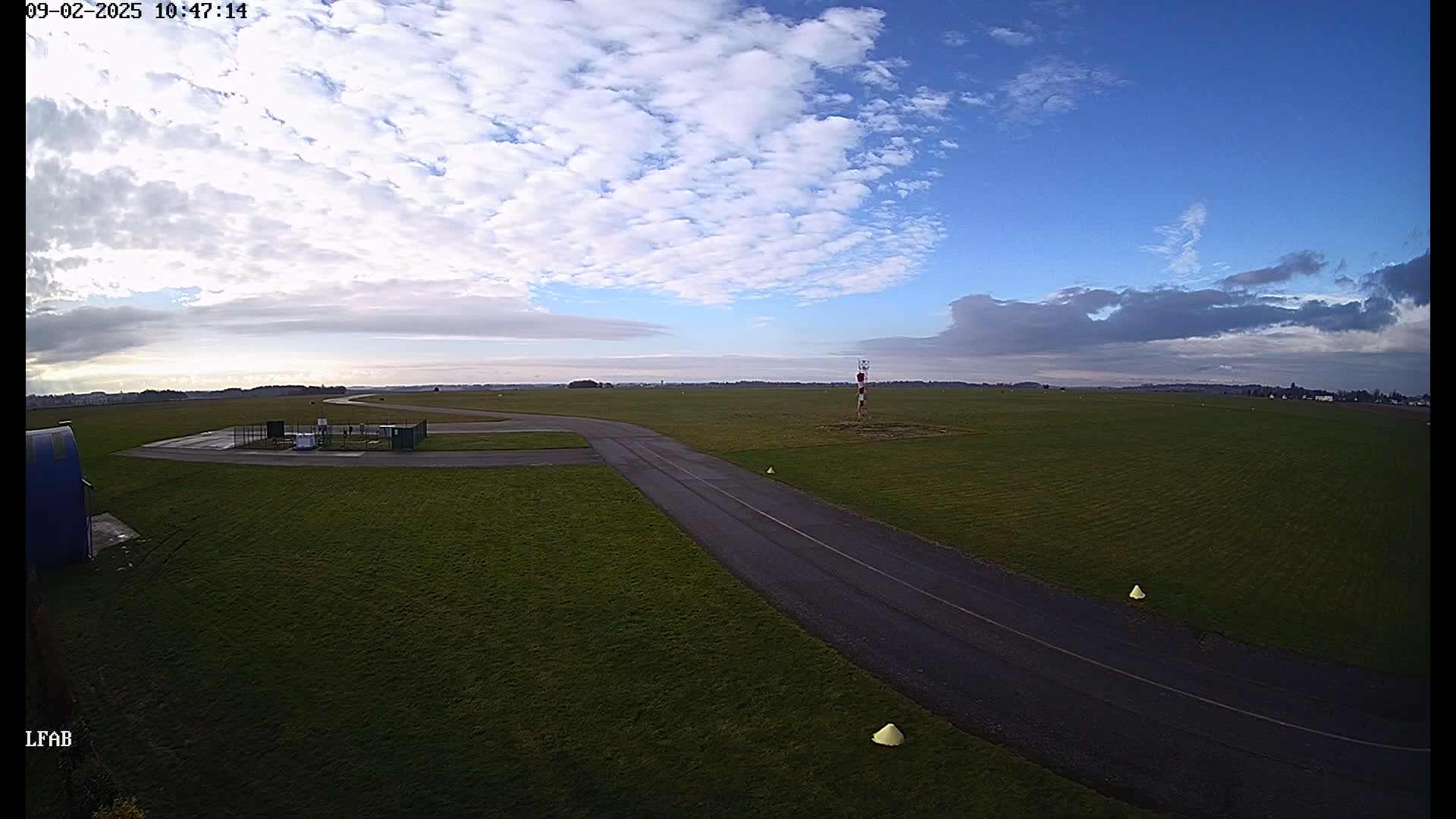 A paved taxiway curves across a grassy airfield under a mostly sunny sky with scattered clouds.