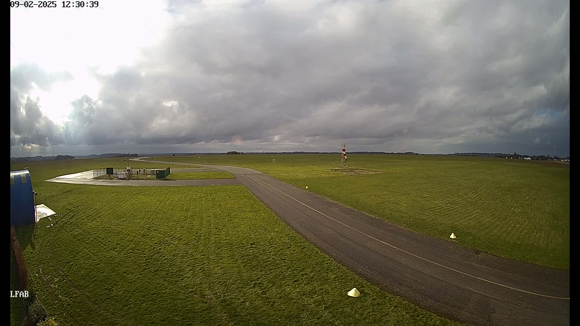 A paved taxiway curves through a grassy airfield under a mostly cloudy sky.