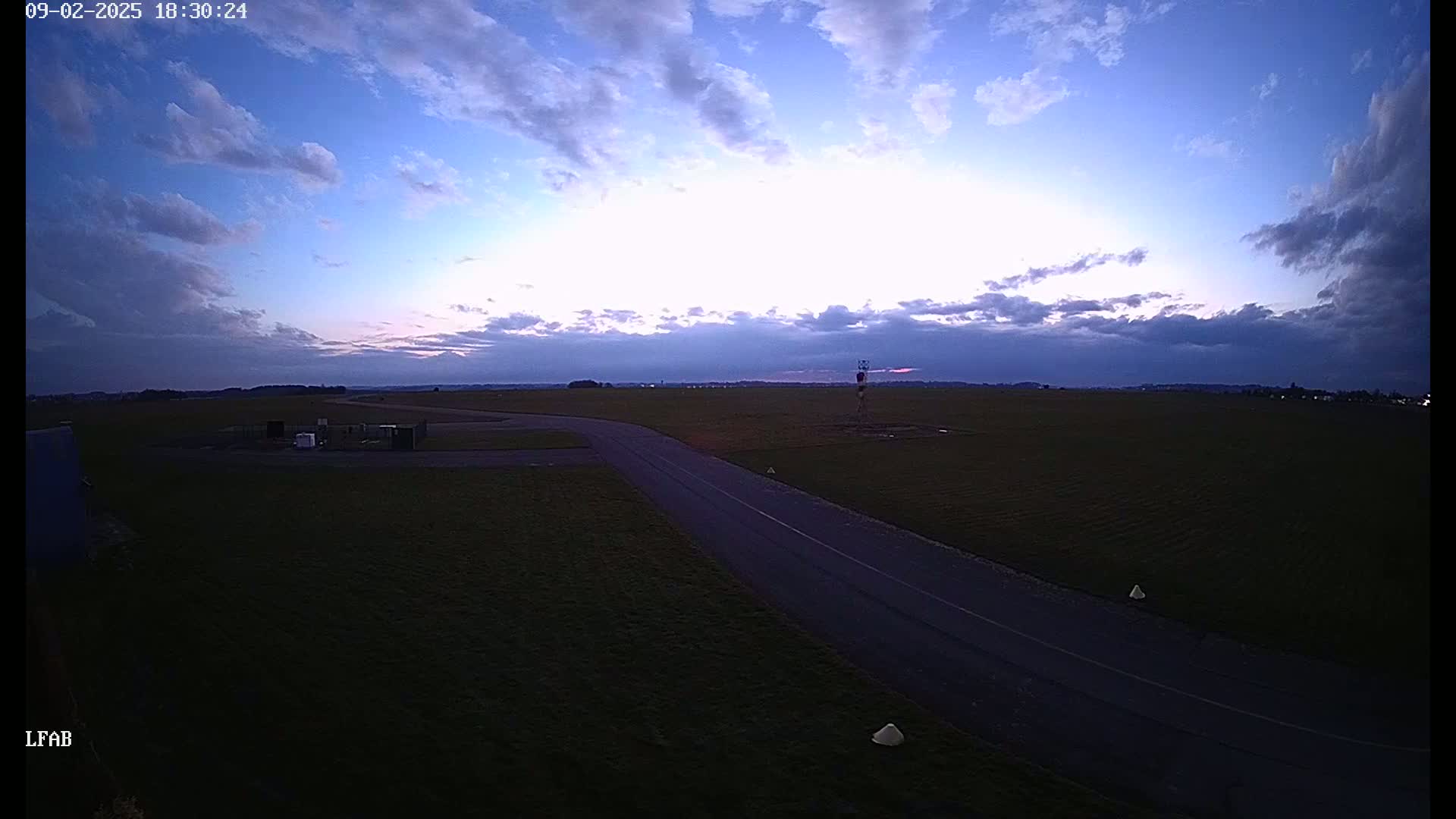 A paved road curves through a grassy airfield at dusk under a partly cloudy sky.