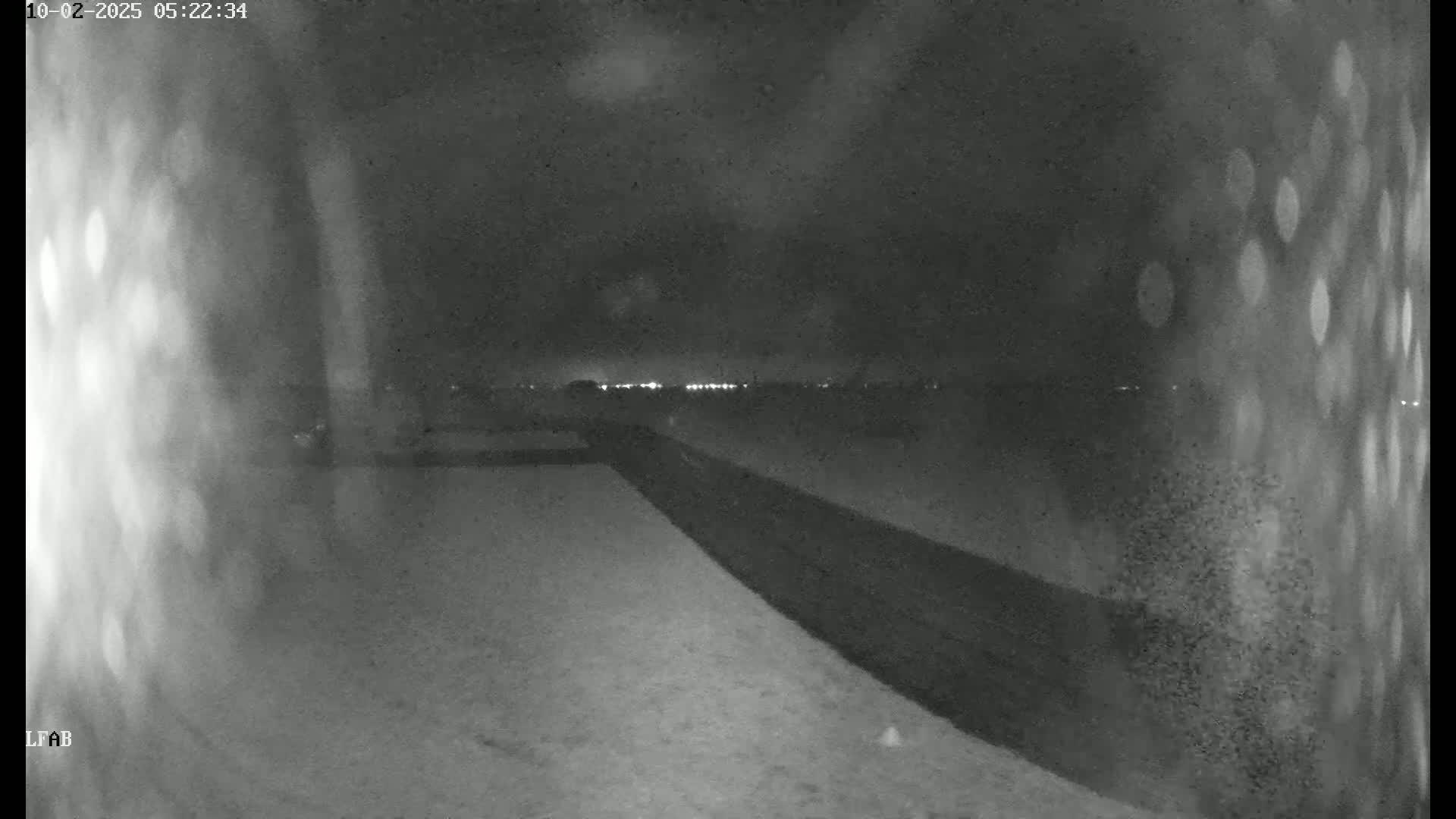 A nighttime, low-light view shows a snow-covered ground with a dark road or embankment leading to a distant line of lights.
