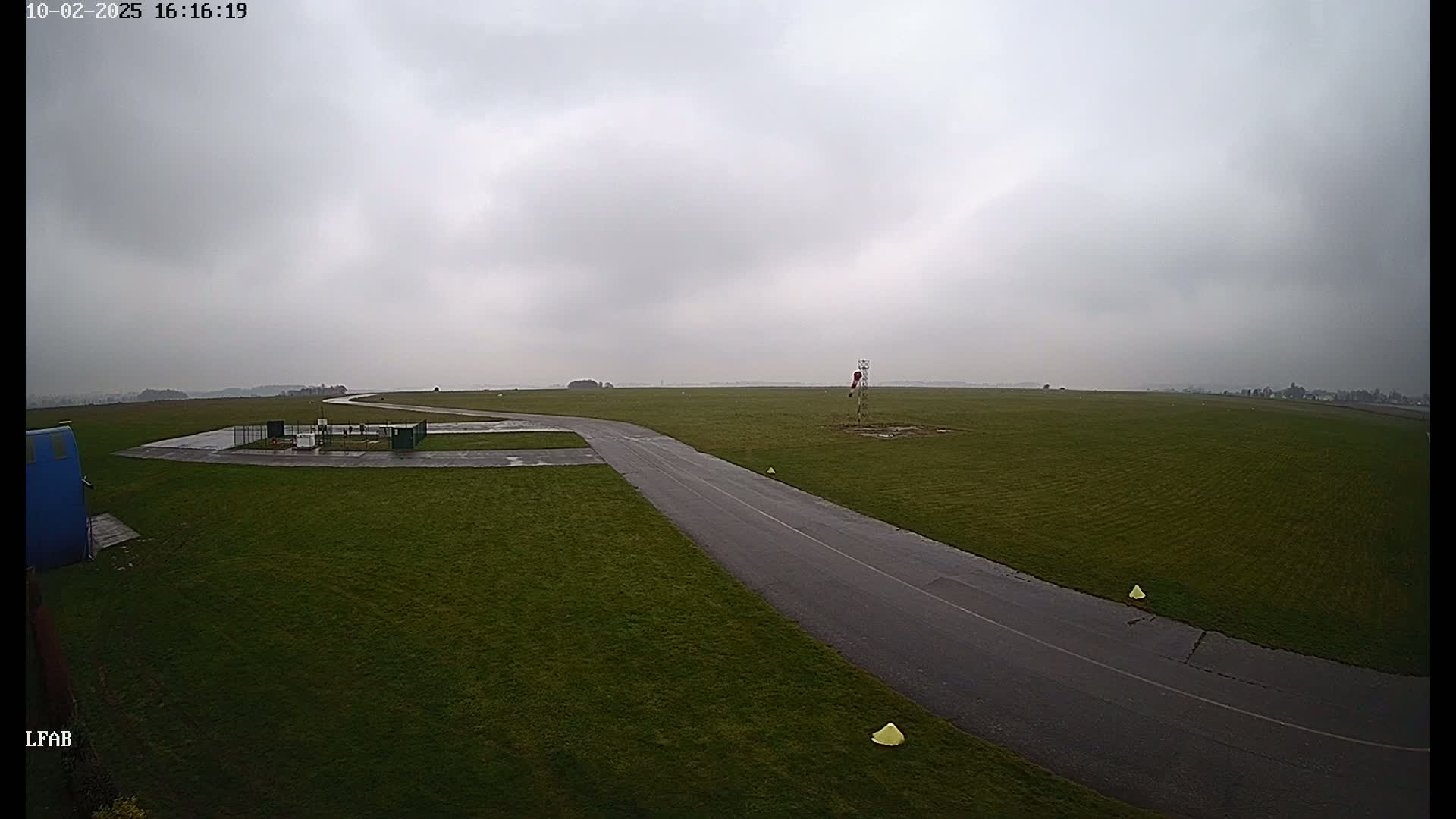 A wet, grassy airfield with a paved taxiway is visible under an overcast sky.