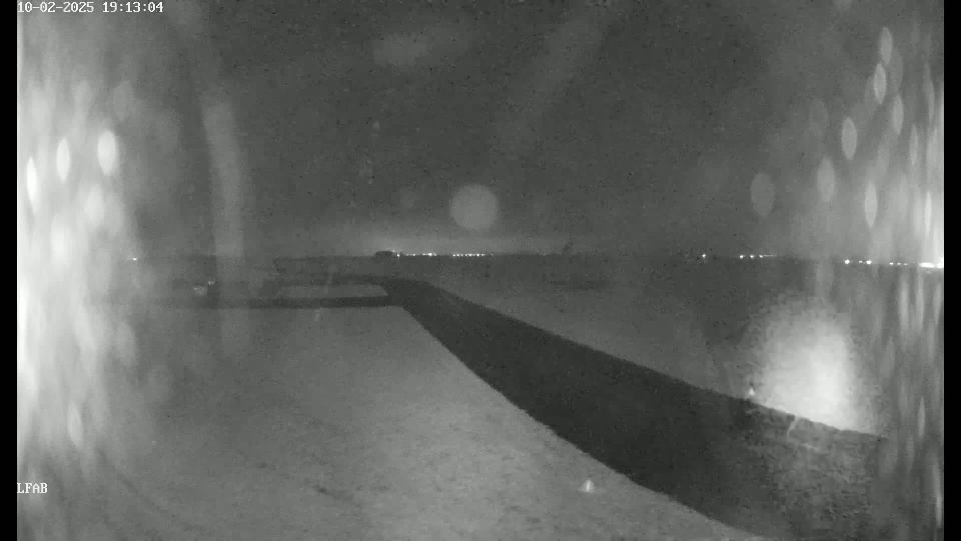 A nighttime, snowy scene shows a dark road stretching into the distance under a cloudy sky with distant lights visible on the horizon.