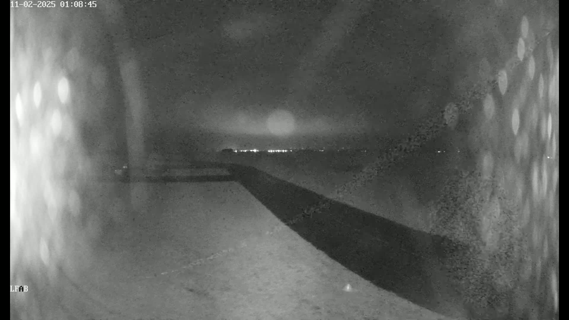 A nighttime, low-light image shows a dark, flat expanse of land, possibly a road or embankment, leading towards a distant line of lights under a cloudy sky.