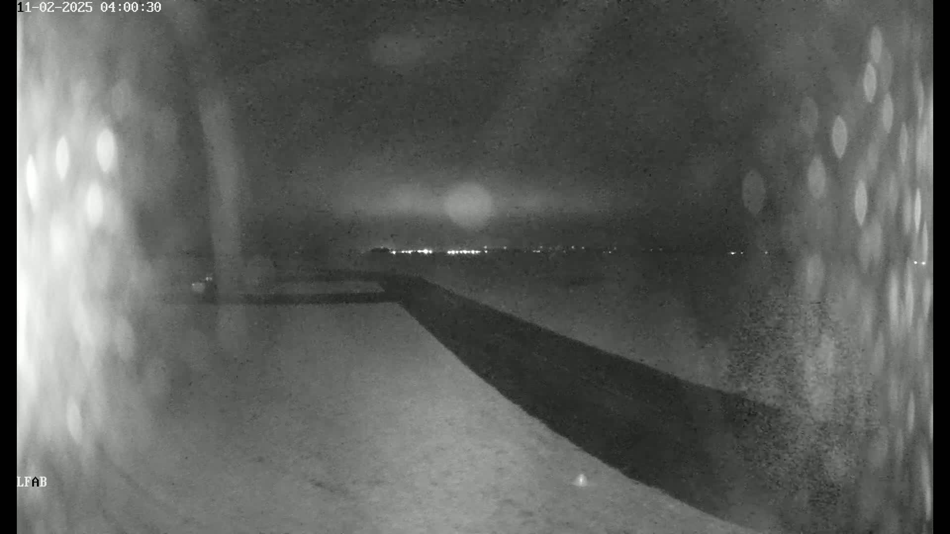 A nighttime, low-light grayscale image shows a snow-covered ground with a dark barrier angled across the frame, and distant lights are visible on the horizon.