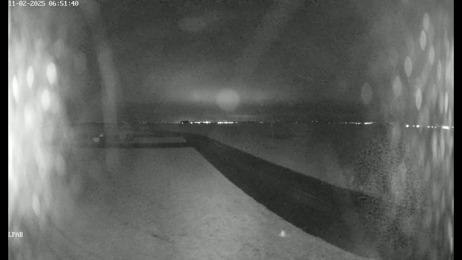 A nighttime, low-light view of a dark road or path leading to a distant horizon with faintly visible lights under a cloudy sky.