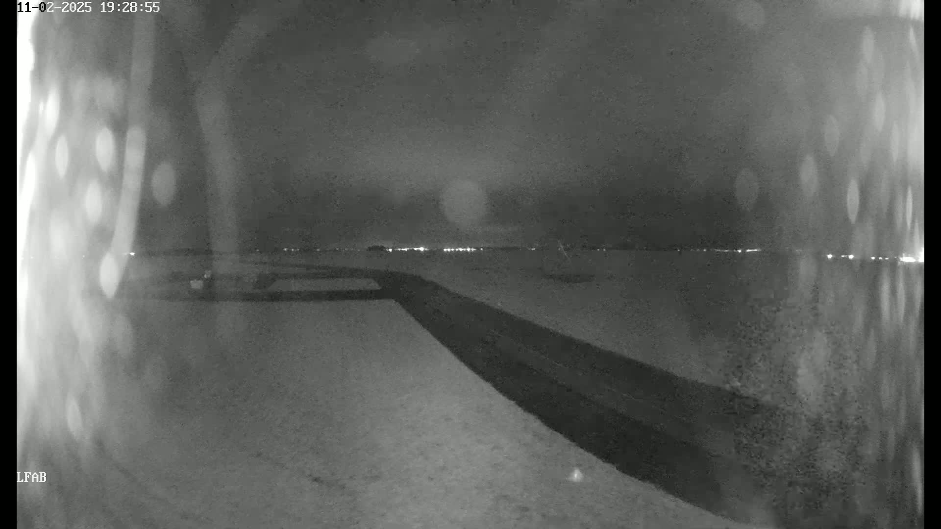 A nighttime, snowy scene shows a dark road curving toward a distant horizon of faint lights under a cloudy sky.