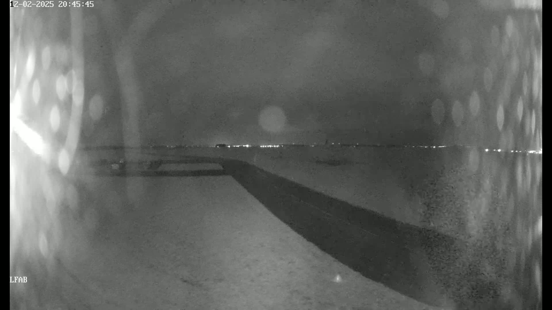 A nighttime, snowy scene shows a dark road leading to a distant line of lights under a cloudy sky.