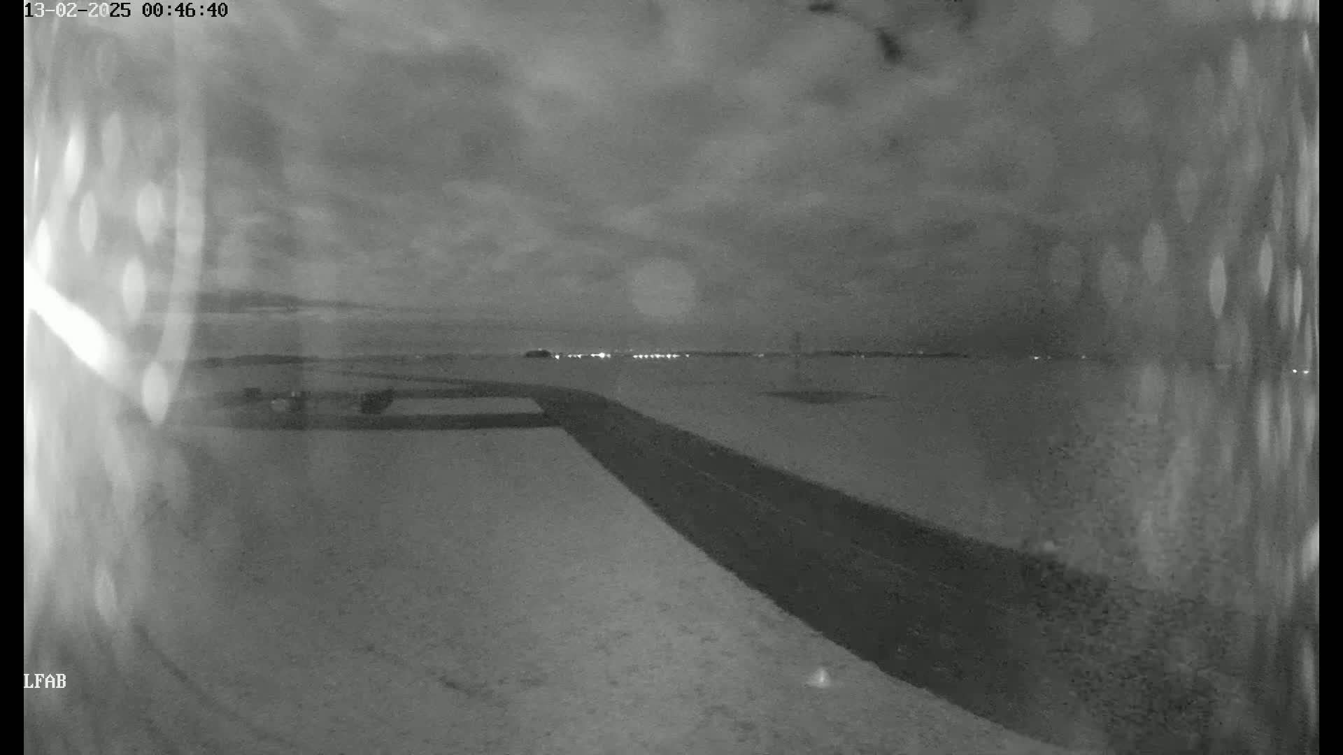 A snow-covered landscape at night is viewed from a high angle, showing a dark road curving toward a distant line of lights under a cloudy sky.