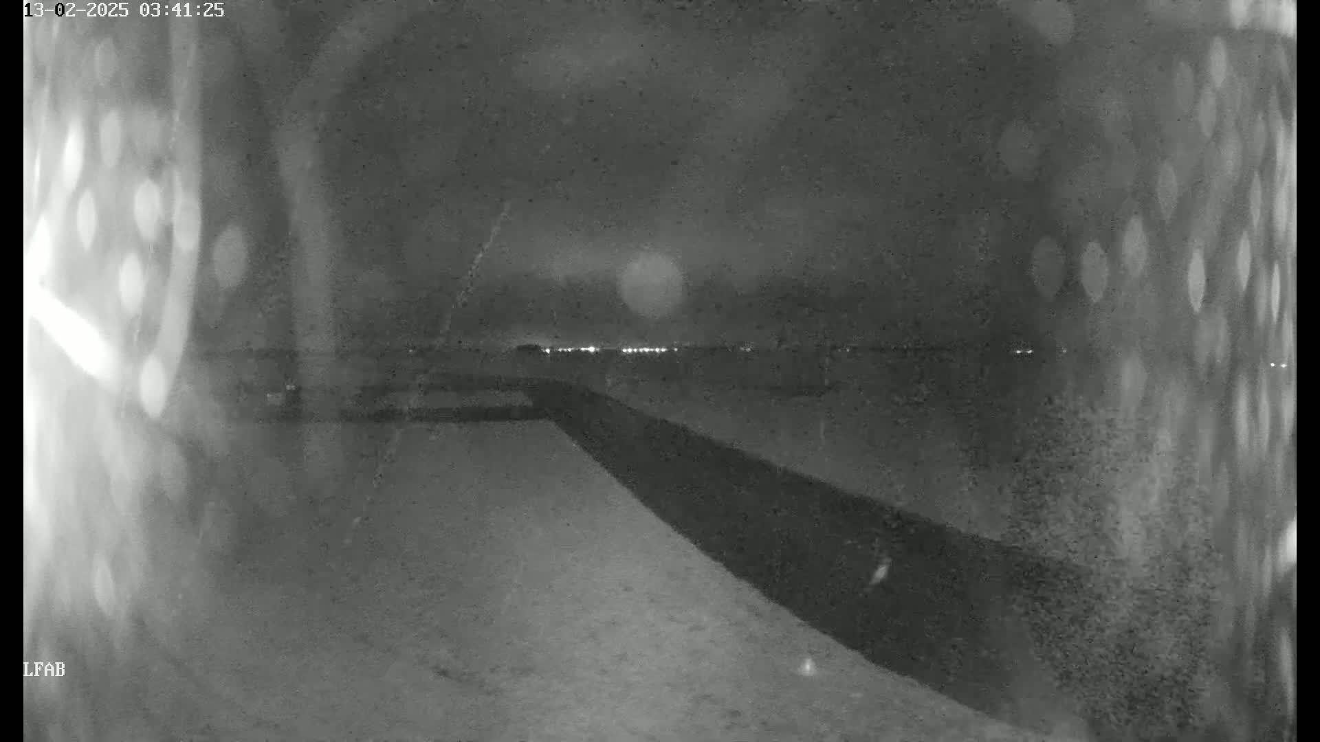 A nighttime, low-light view shows a snow-covered area with a dark barrier extending from the foreground toward a distant horizon dotted with faint lights.