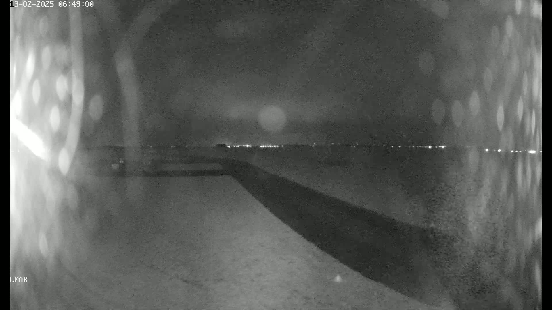 A nighttime, low-light view shows a flat, dark expanse with a distant line of lights and a dark structure extending from the bottom right to the middle of the image.