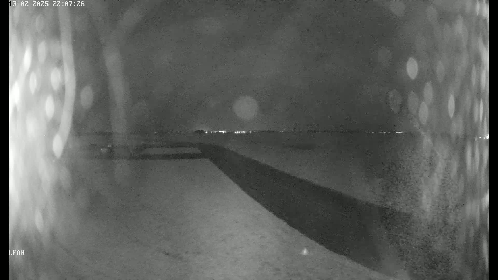A nighttime, snowy scene shows a dark road or path leading to a distant line of lights under a cloudy sky.