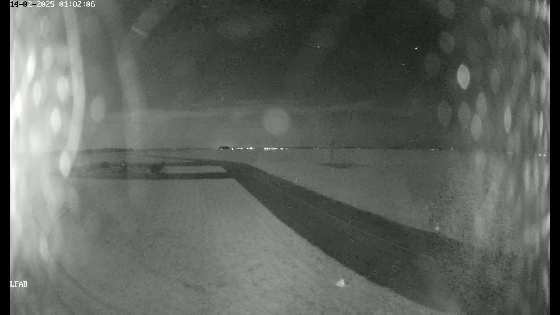 A snow-covered road at night is seen from a low angle, with a distant line of lights visible on the horizon.