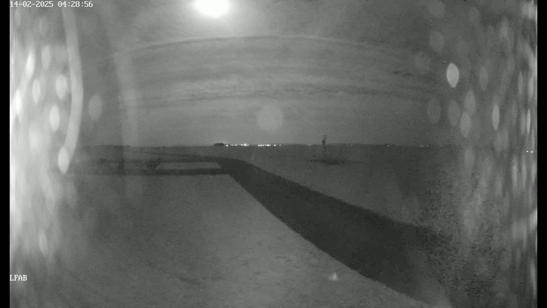 A nighttime, low-light view shows a dark, paved area extending toward a horizon where faint lights are visible under a bright moon and cloudy sky.