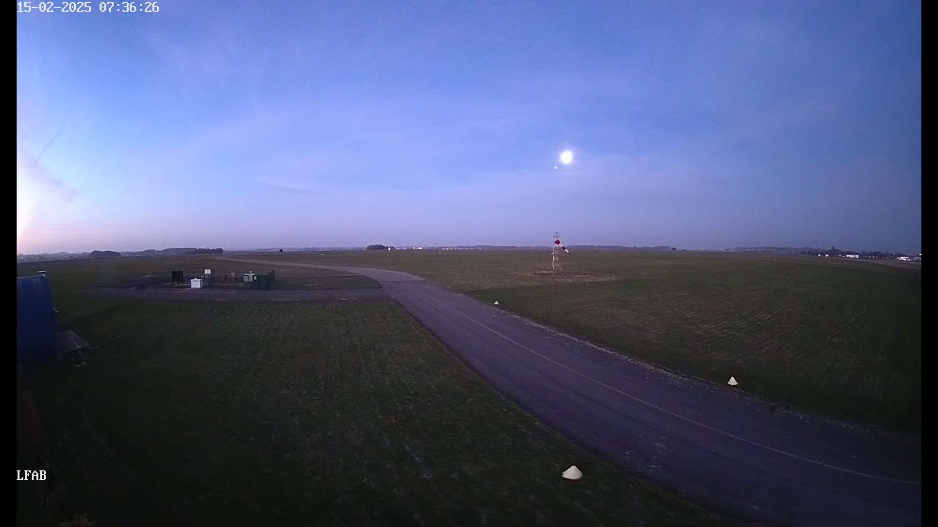 A paved road curves through a flat, grassy field under a twilight sky with a bright moon visible.