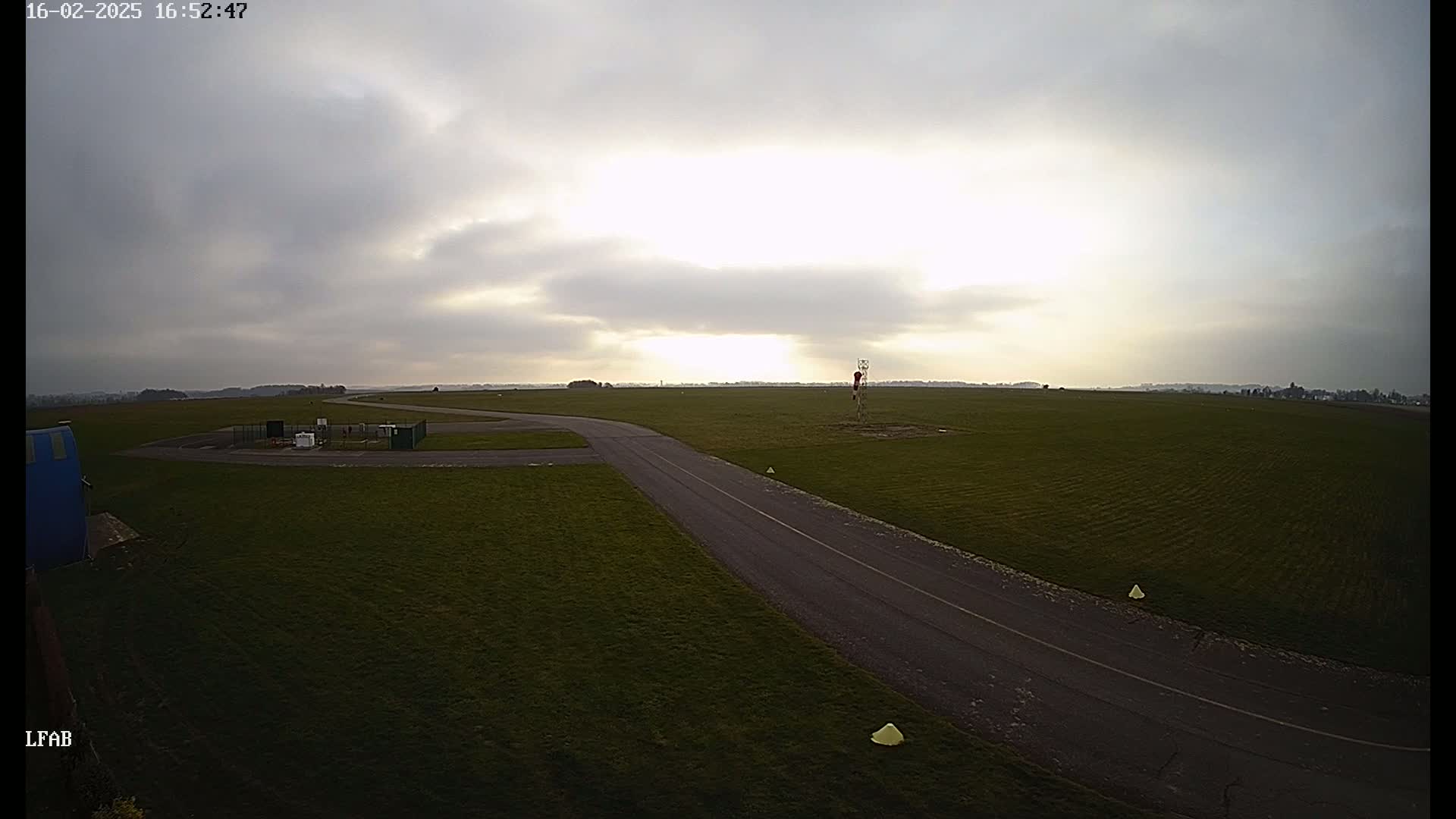 A paved taxiway curves through a grassy airfield under a mostly cloudy sky, with the sun partially visible.