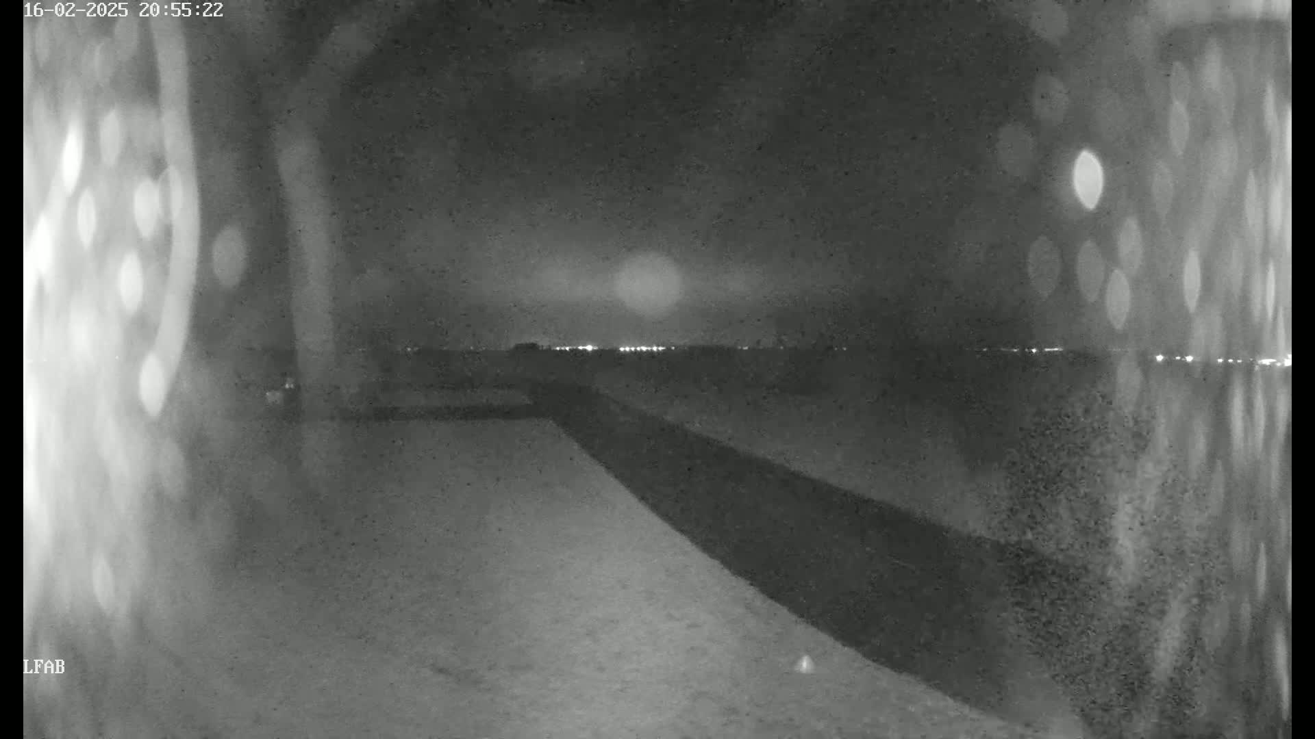 A nighttime, blurry view shows a dark road extending into a flat, snow-covered landscape under a cloudy sky with a bright light source in the distance.