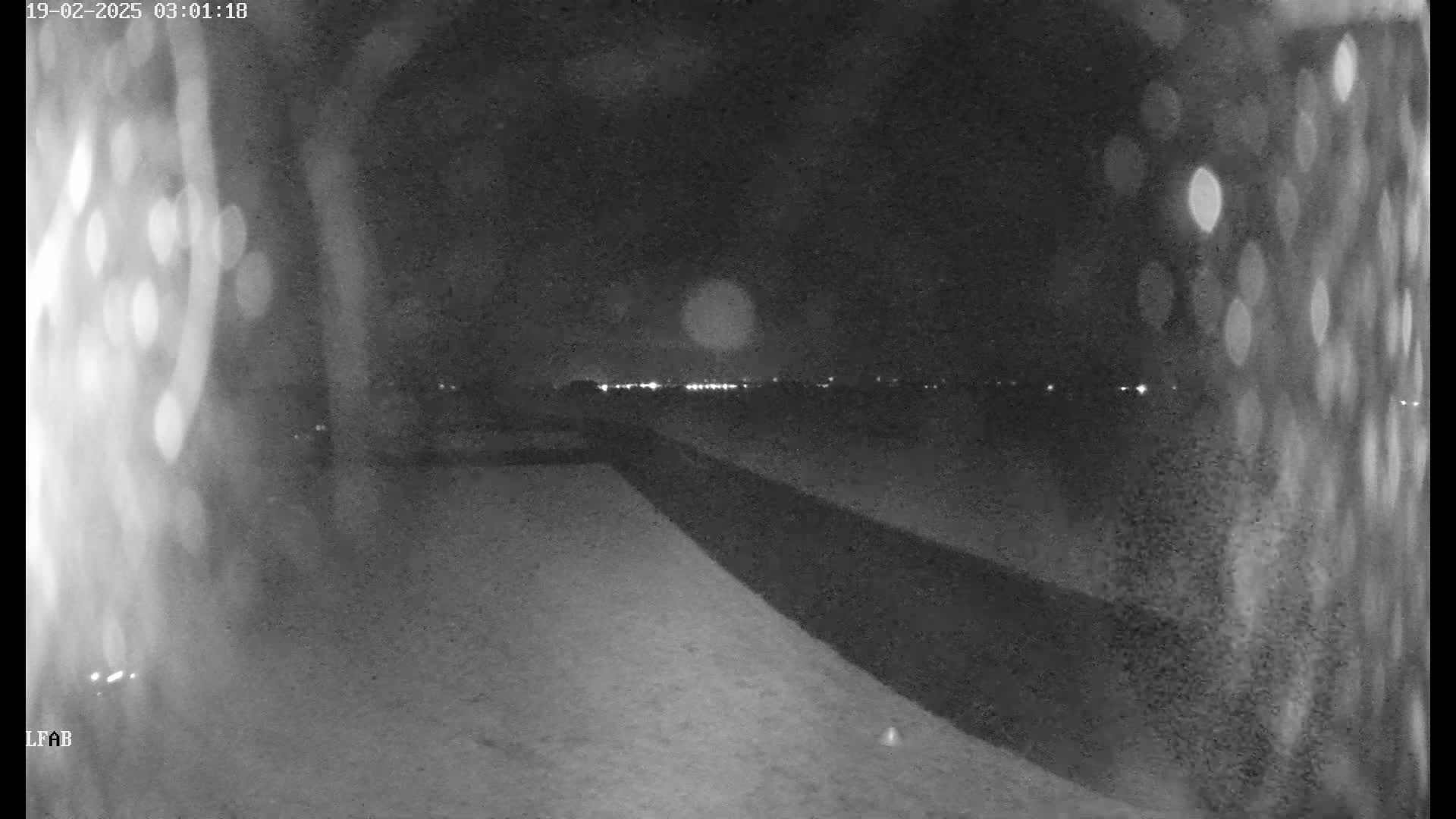 A nighttime, low-light view shows a dark, flat expanse of land leading to a line of distant lights under a mostly clear sky.