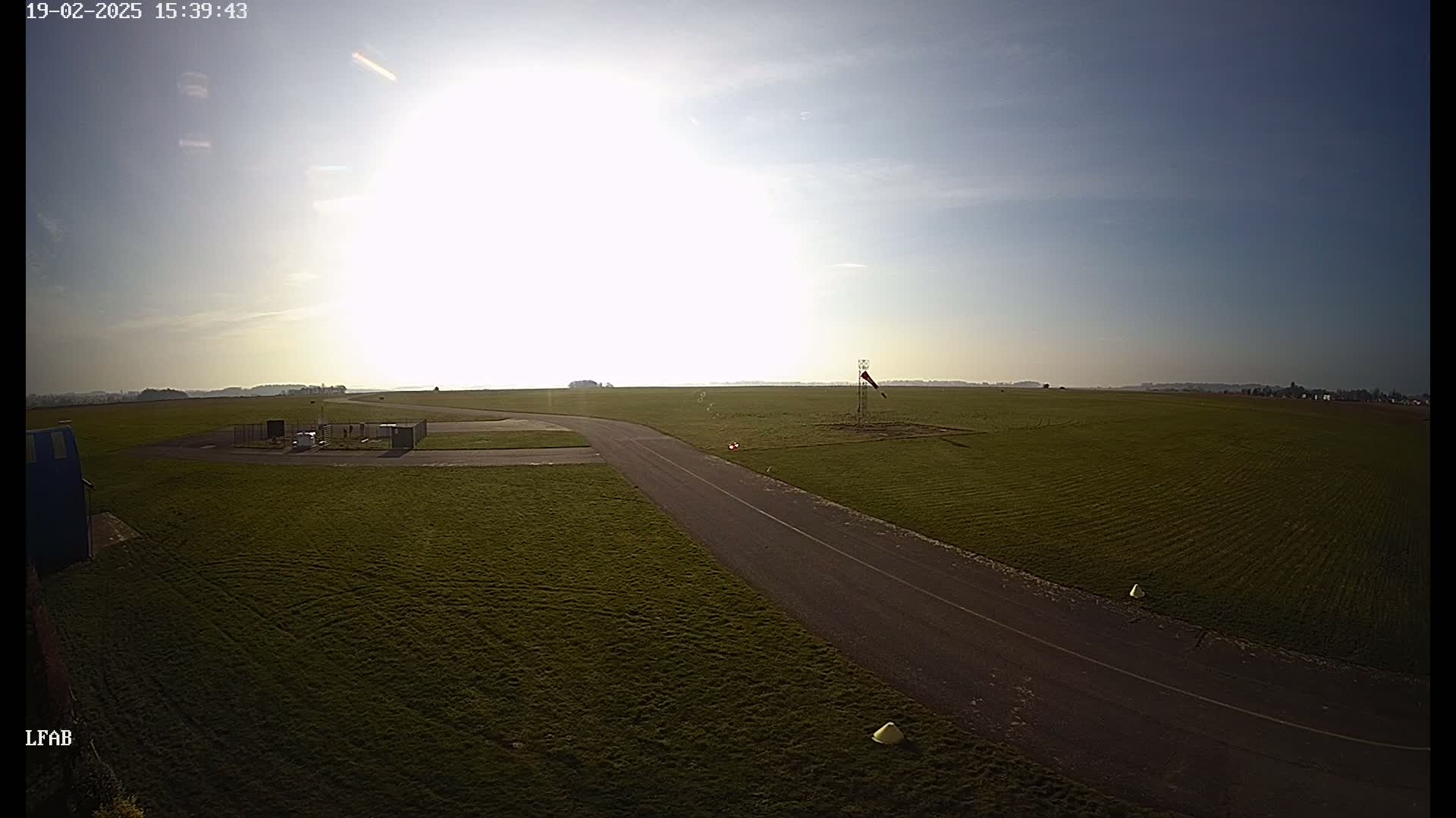 A sunny day at a grass airfield shows a paved taxiway leading to a grassy field, with a windsock visible in the distance.