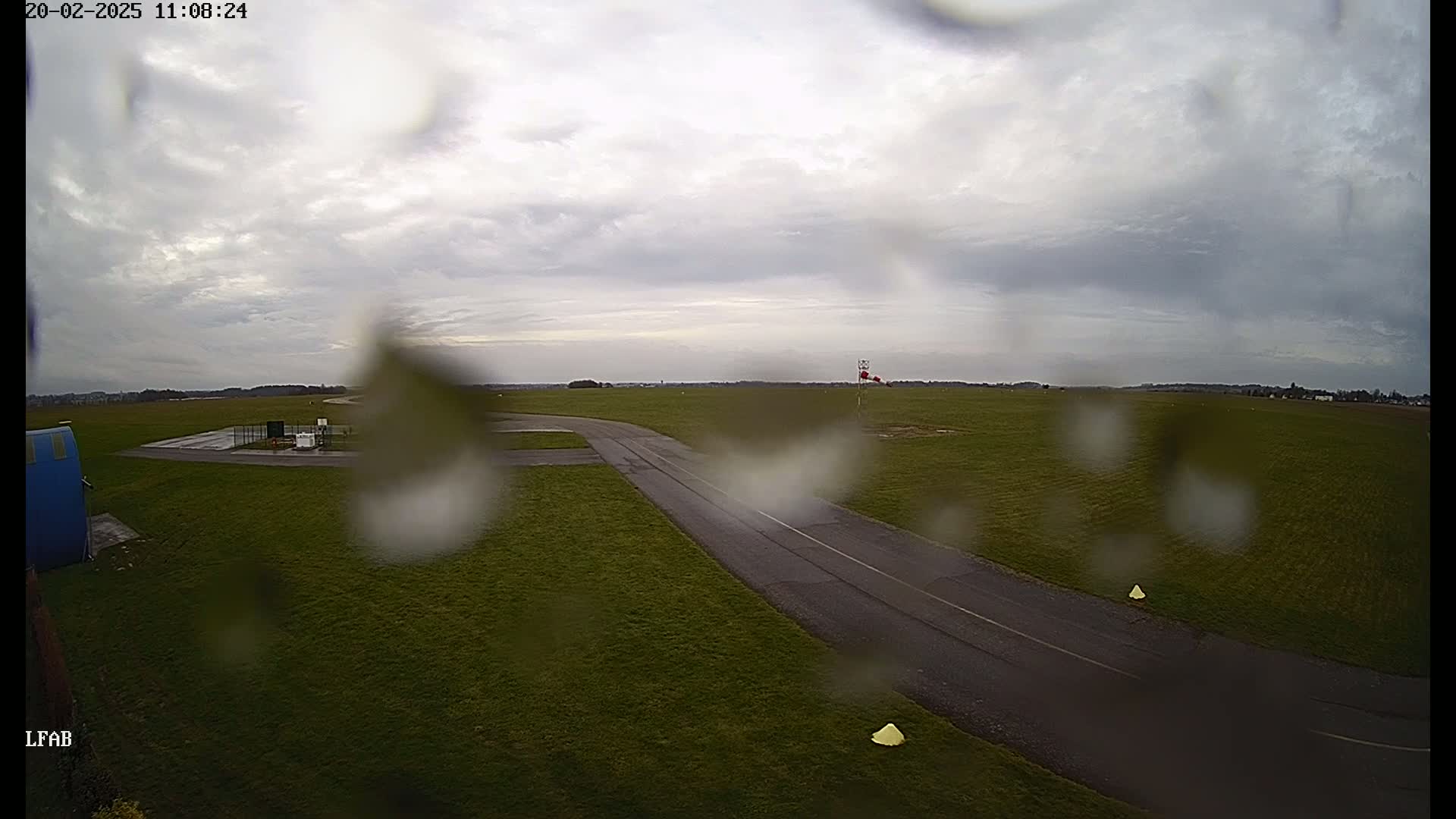 A rain-covered view of a grassy airfield with a paved taxiway, under a cloudy sky.