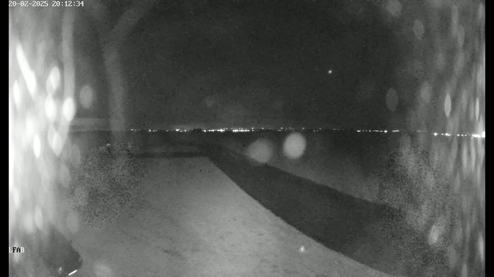 A nighttime, low-light view shows a dark, sloped surface leading to a distant horizon with faint city lights under a clear sky.