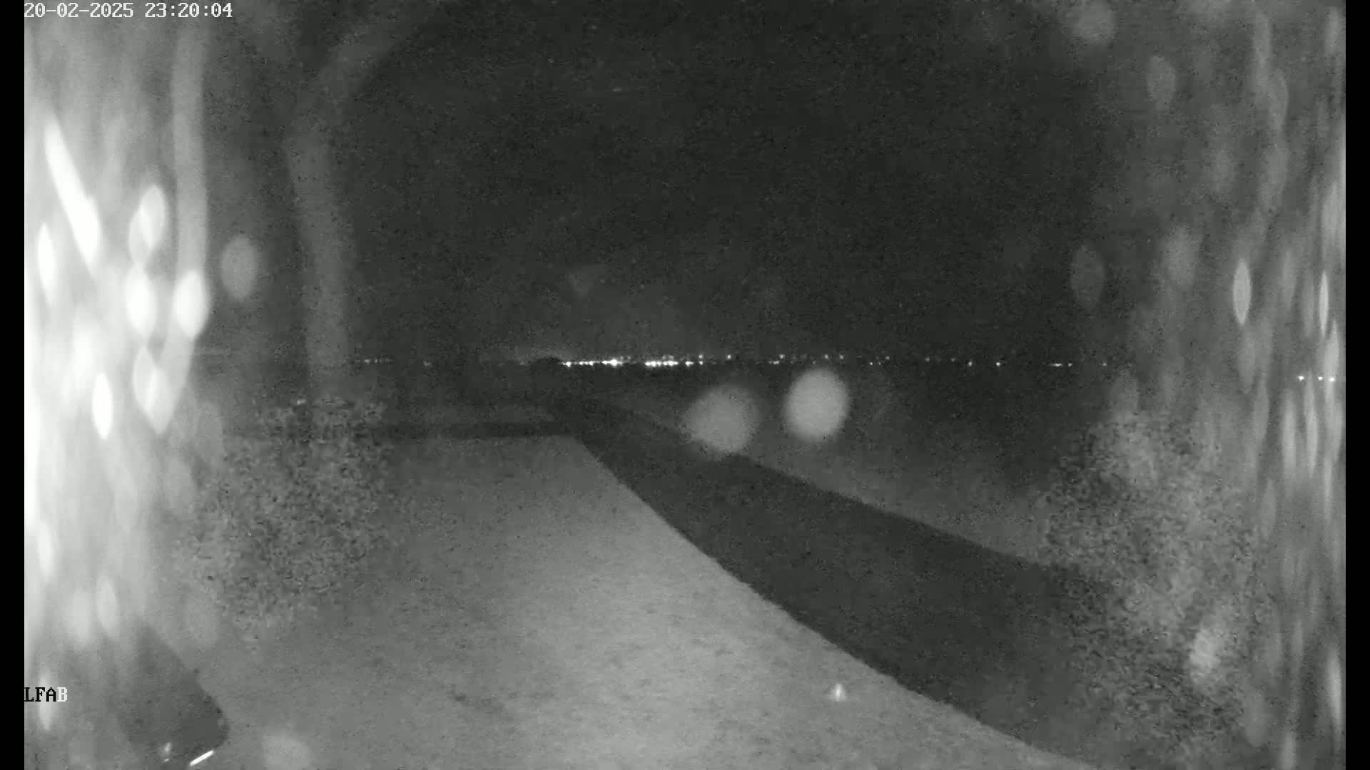 A nighttime, blurry outdoor view shows a dark paved area sloping down towards a distant line of lights.