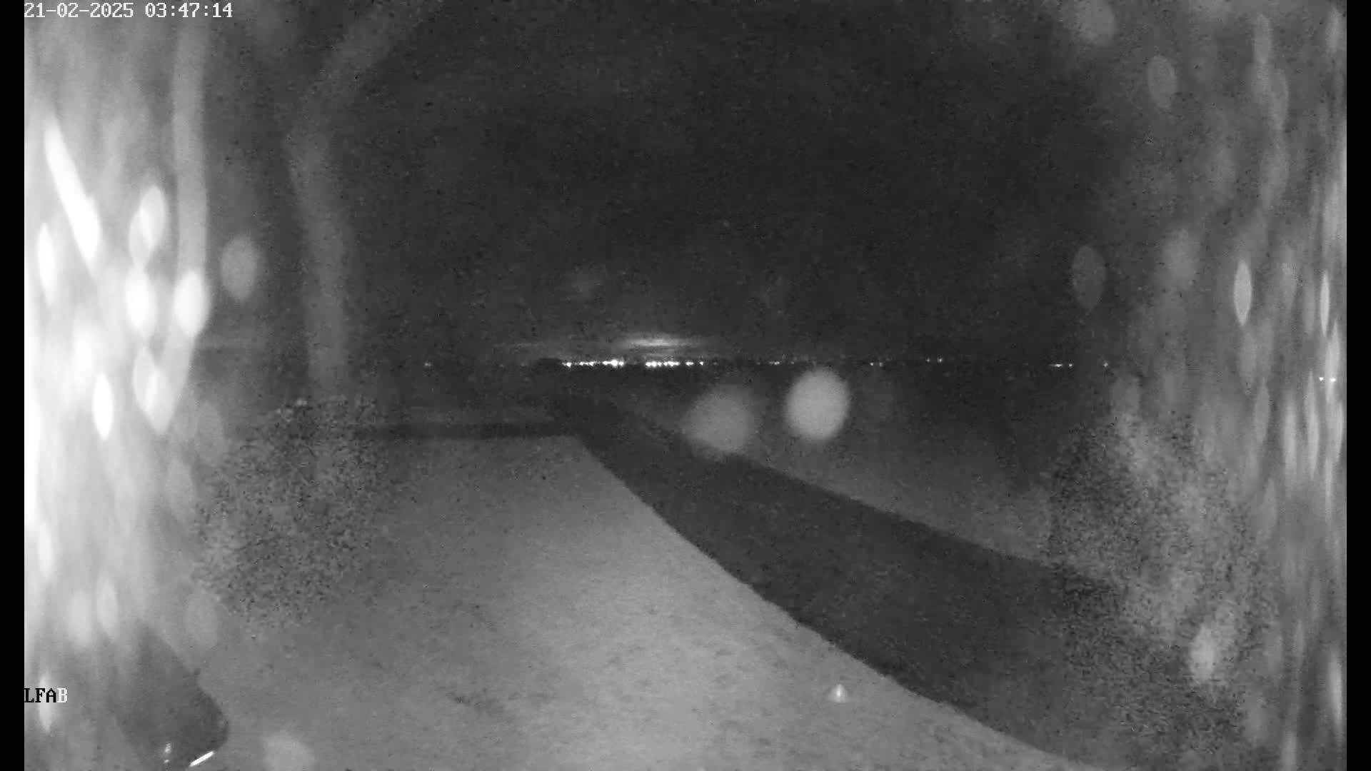 A nighttime, blurry view shows a dark, snow-covered ground sloping down towards a distant line of lights under a dark sky.