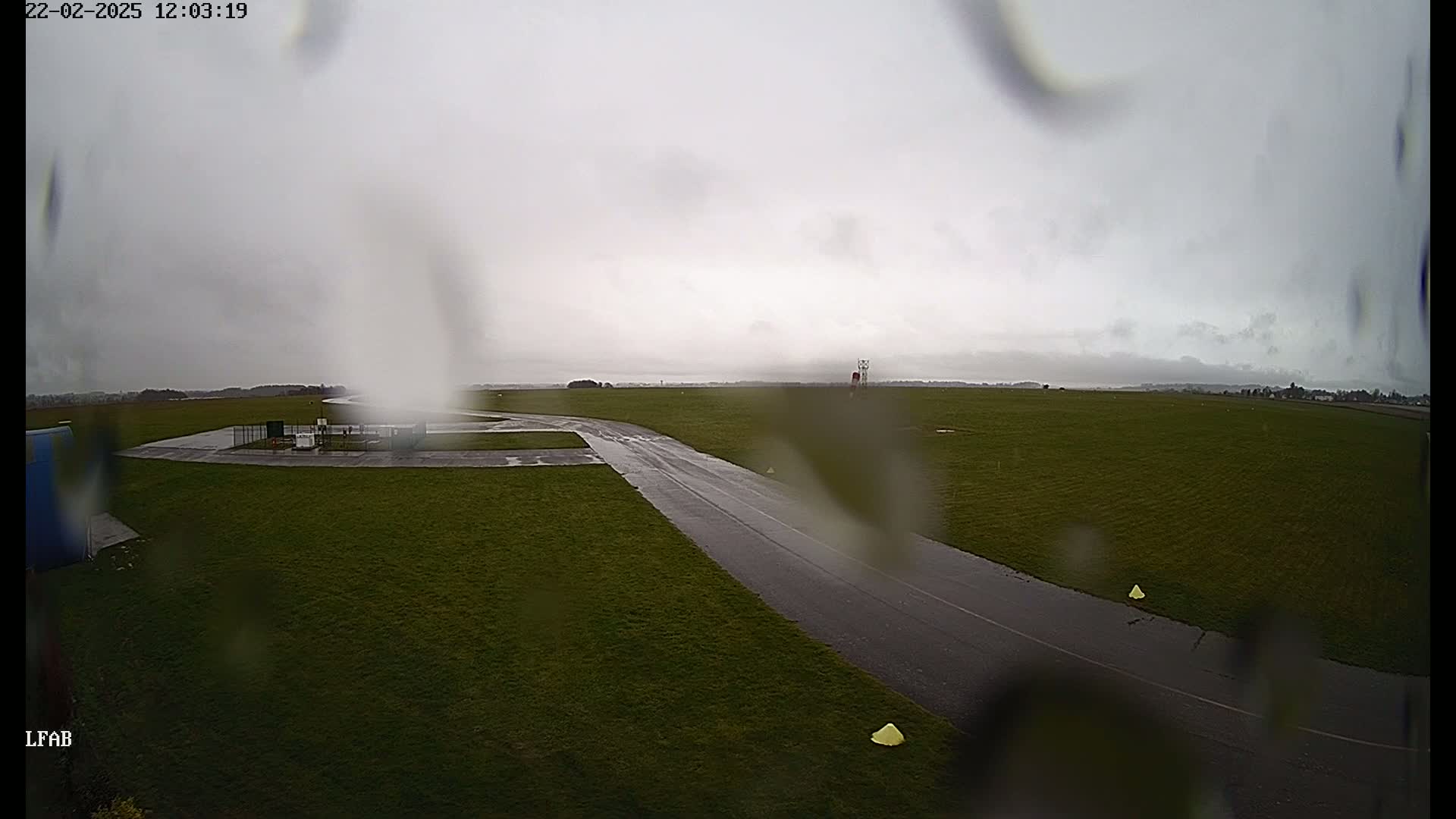 A wet, paved taxiway curves through a grassy airfield under an overcast sky during rainy weather.