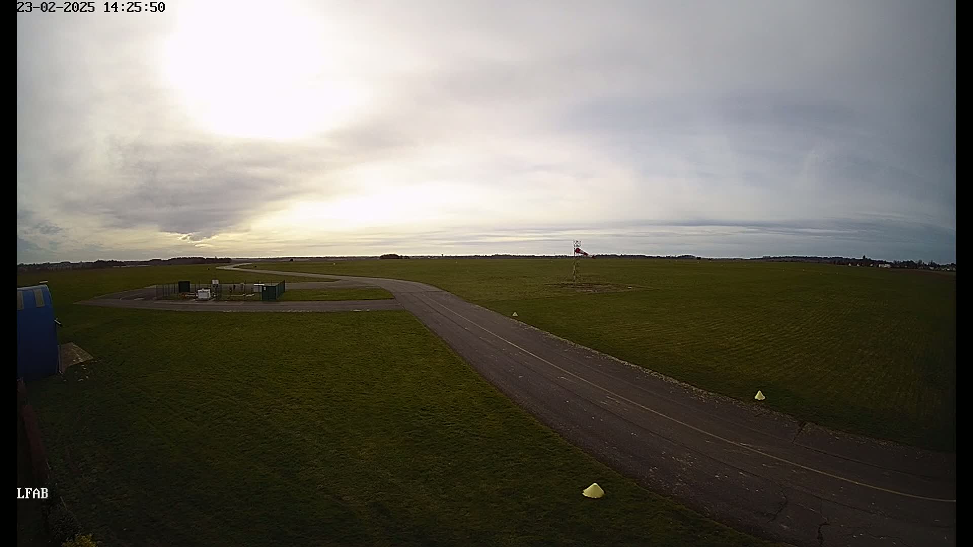 An overcast sky hangs over a grassy airfield with a paved taxiway and windsock.