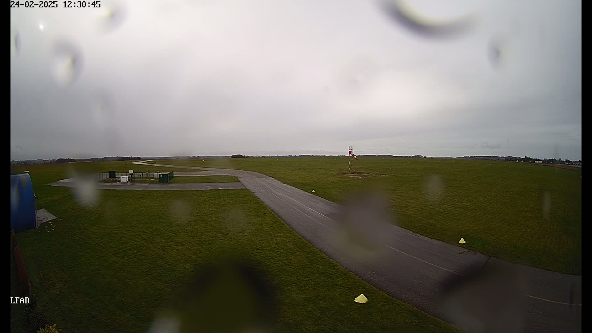 A rain-covered view of a grassy airfield with a paved taxiway, a windsock, and some small markers.