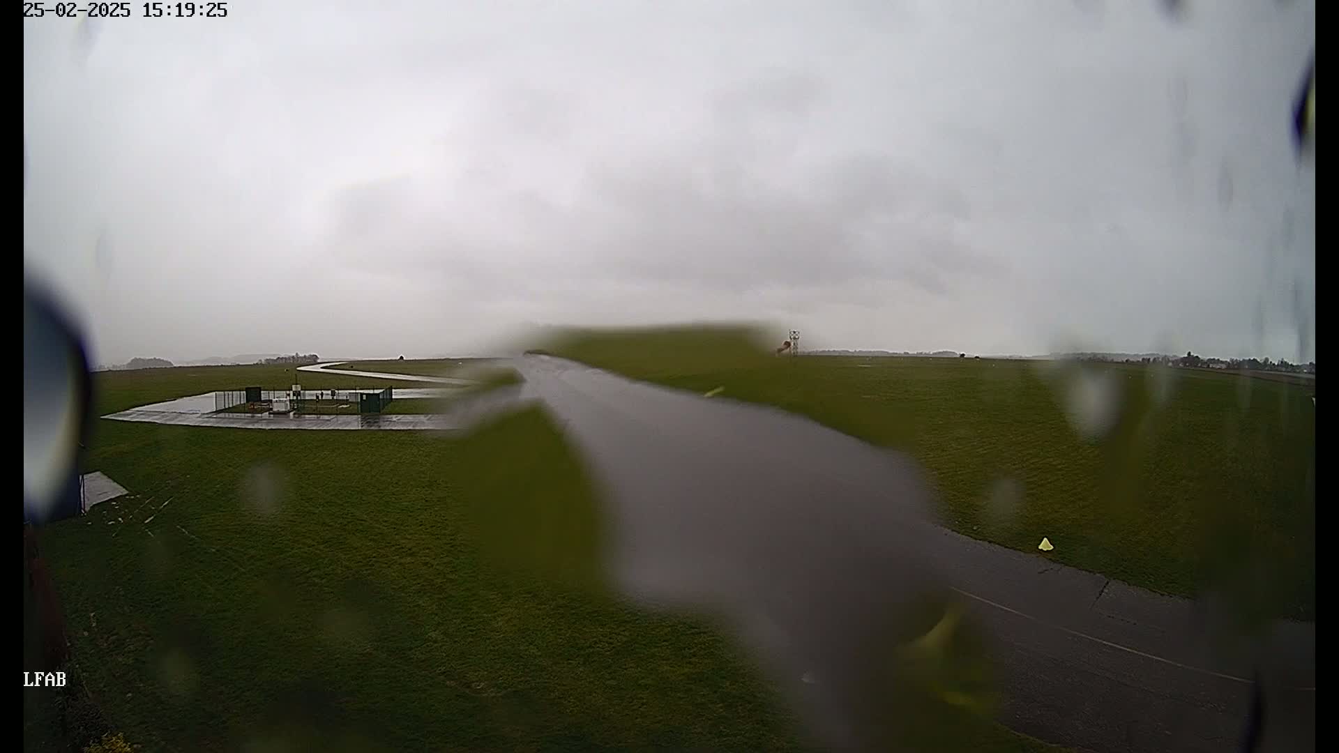 A rain-streaked view shows a grassy airfield with a paved runway and small structures under an overcast sky.