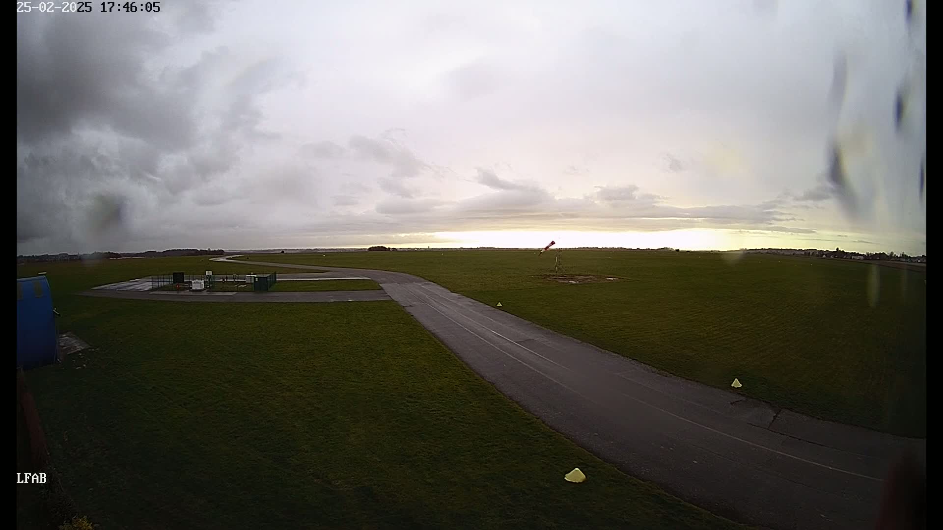 A tarmac taxiway winds across a grassy airfield under a mostly cloudy, overcast sky.