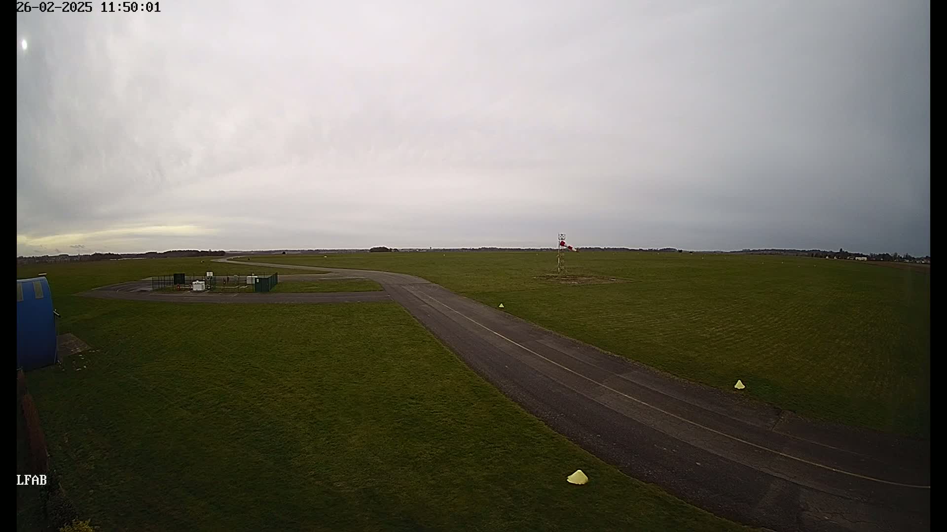 An overcast sky hangs over a grassy airfield with a paved taxiway leading to a small building.
