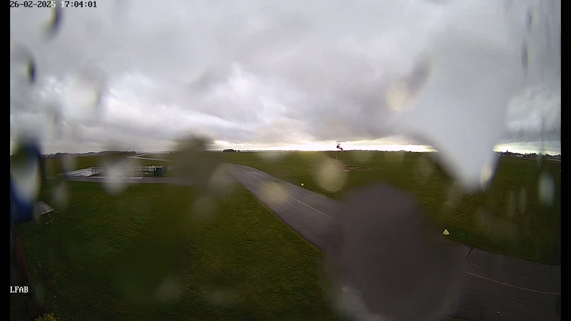 A rain-streaked view shows a grassy airfield with a paved runway under an overcast sky.